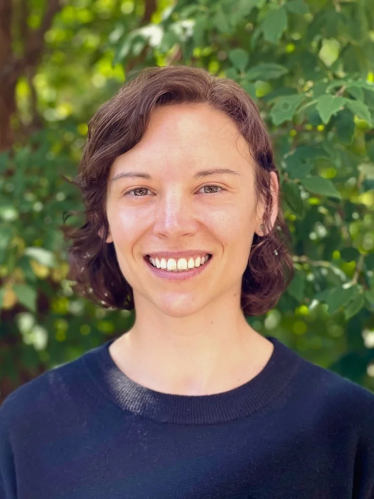 A young woman with short, curly brown hair smiling outdoors in front of green leafy foliage.