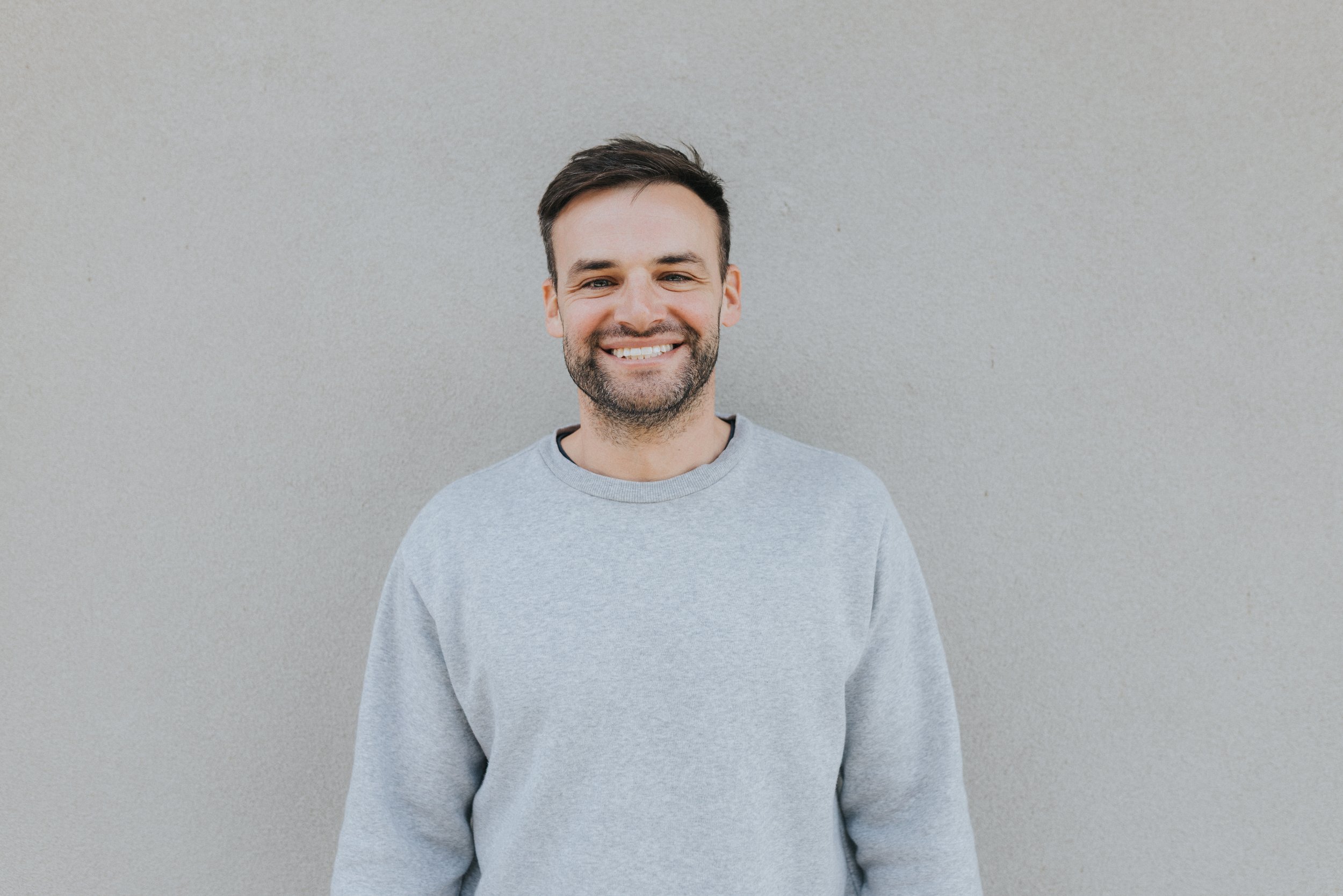 Smiling man with dark hair and beard wearing a light gray sweatshirt standing against a plain light gray wall.