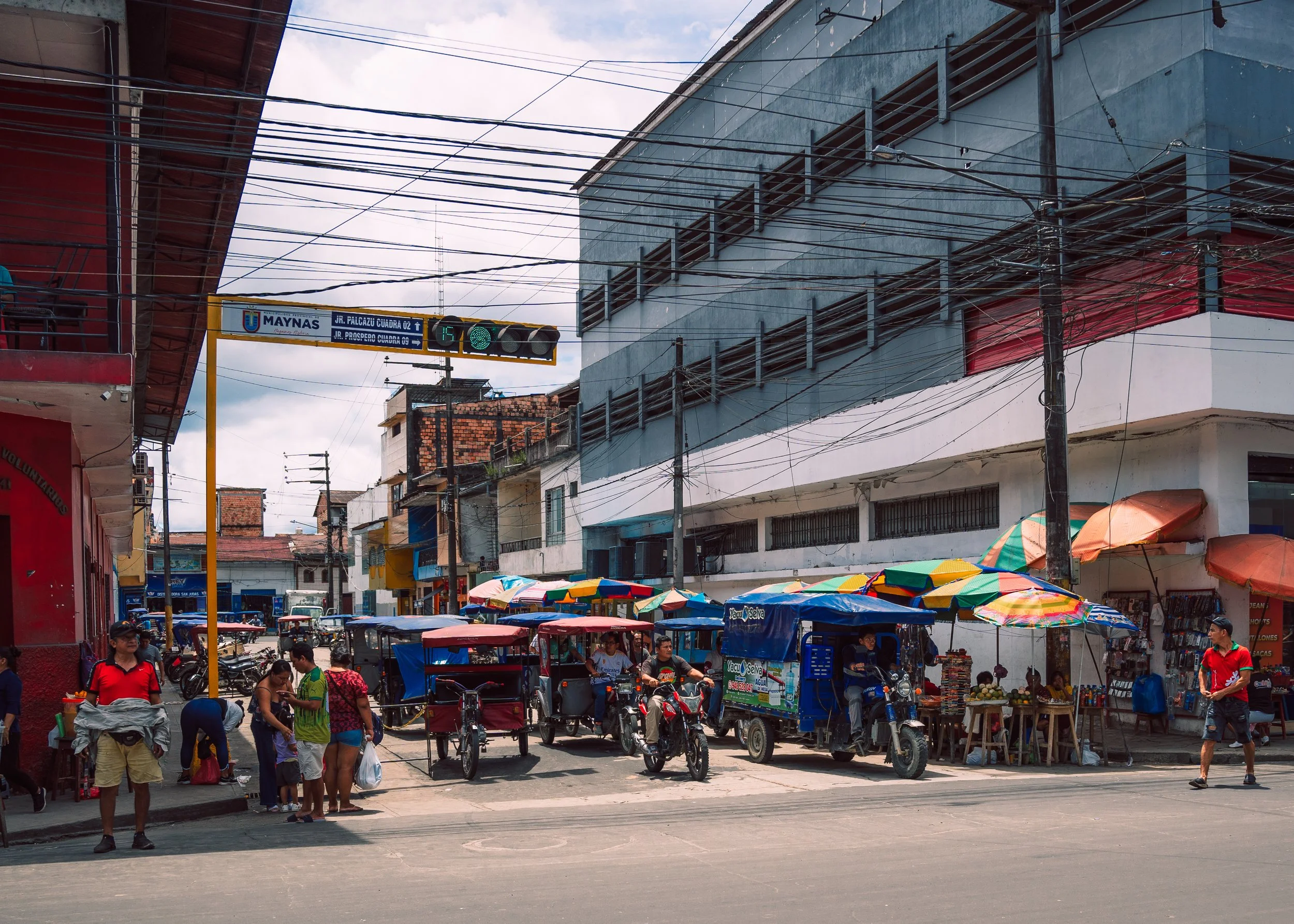 Street market scene with vendors on motorcycles and tricycles, colorful umbrellas, and pedestrians on a busy city street.
