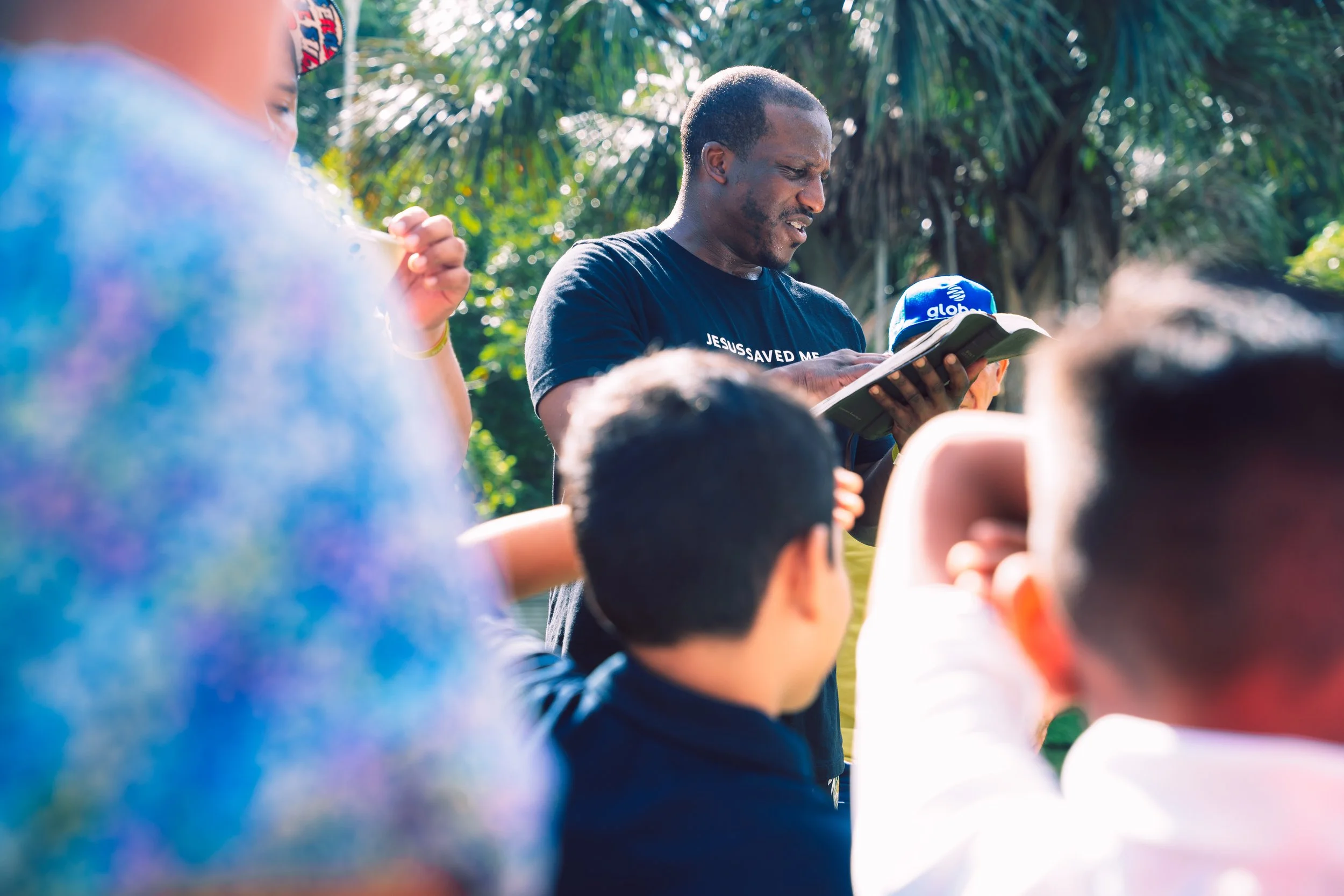 Man leading outdoor religious service with children gathered around, reading from a book, in a tropical setting.
