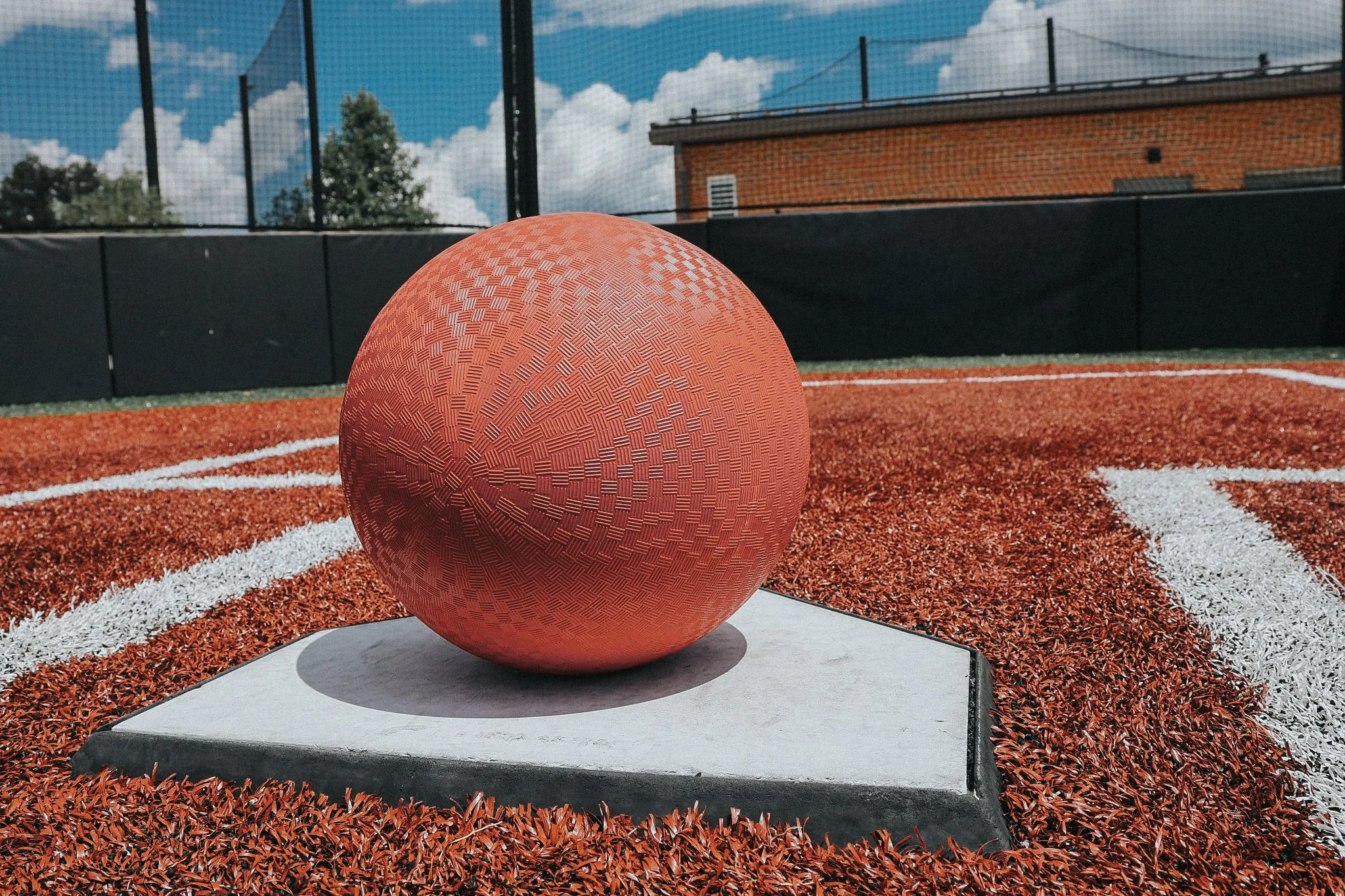 Close-up of a textured red basketball on a sports court with white lines, surrounded by red and white turf, with a black fence and a building in the background under a partly cloudy sky.