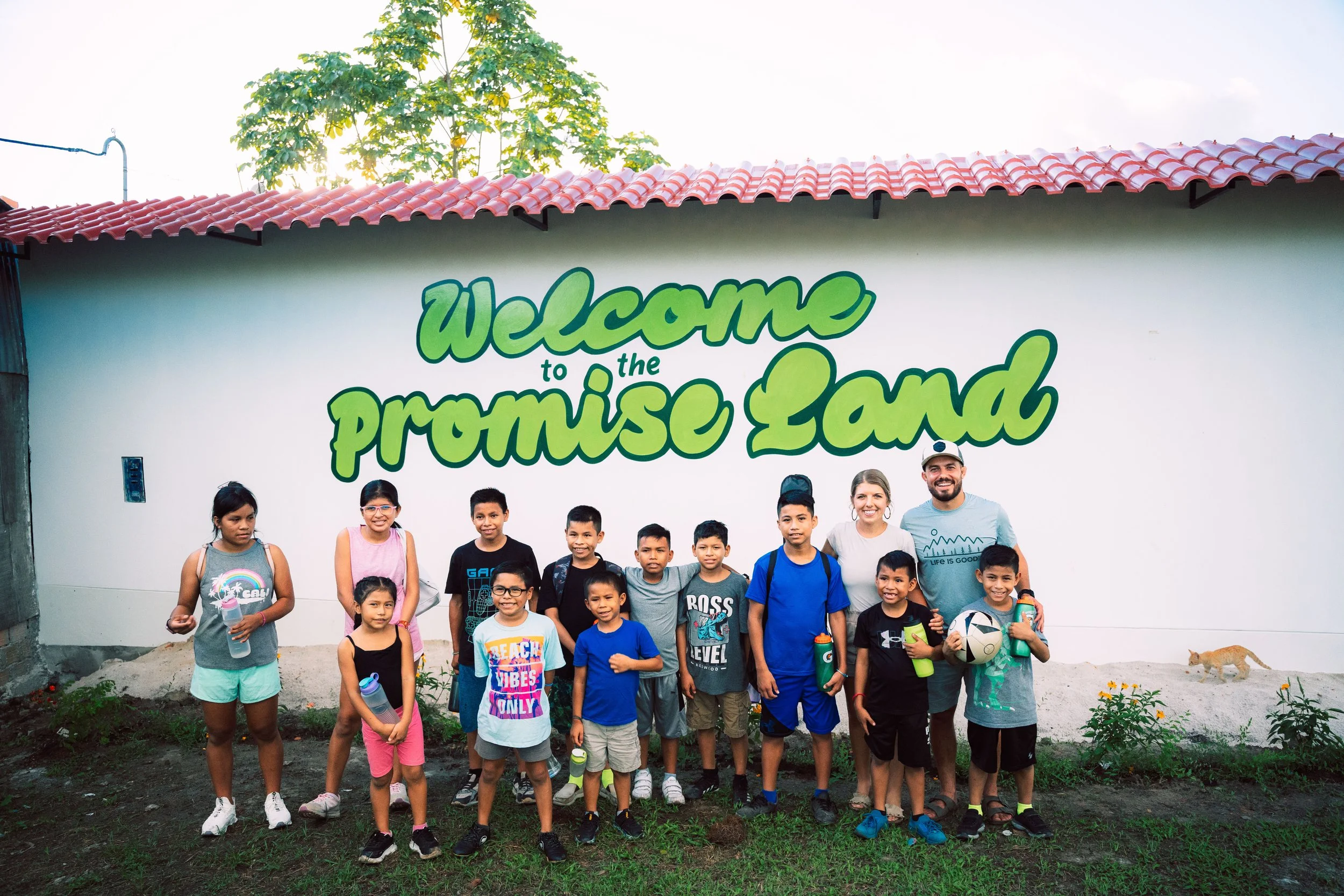 Group of children and two adults standing outside in front of a white wall with a green sign that reads 'Welcome to the Promise Land.' Some children hold water bottles and sports equipment.