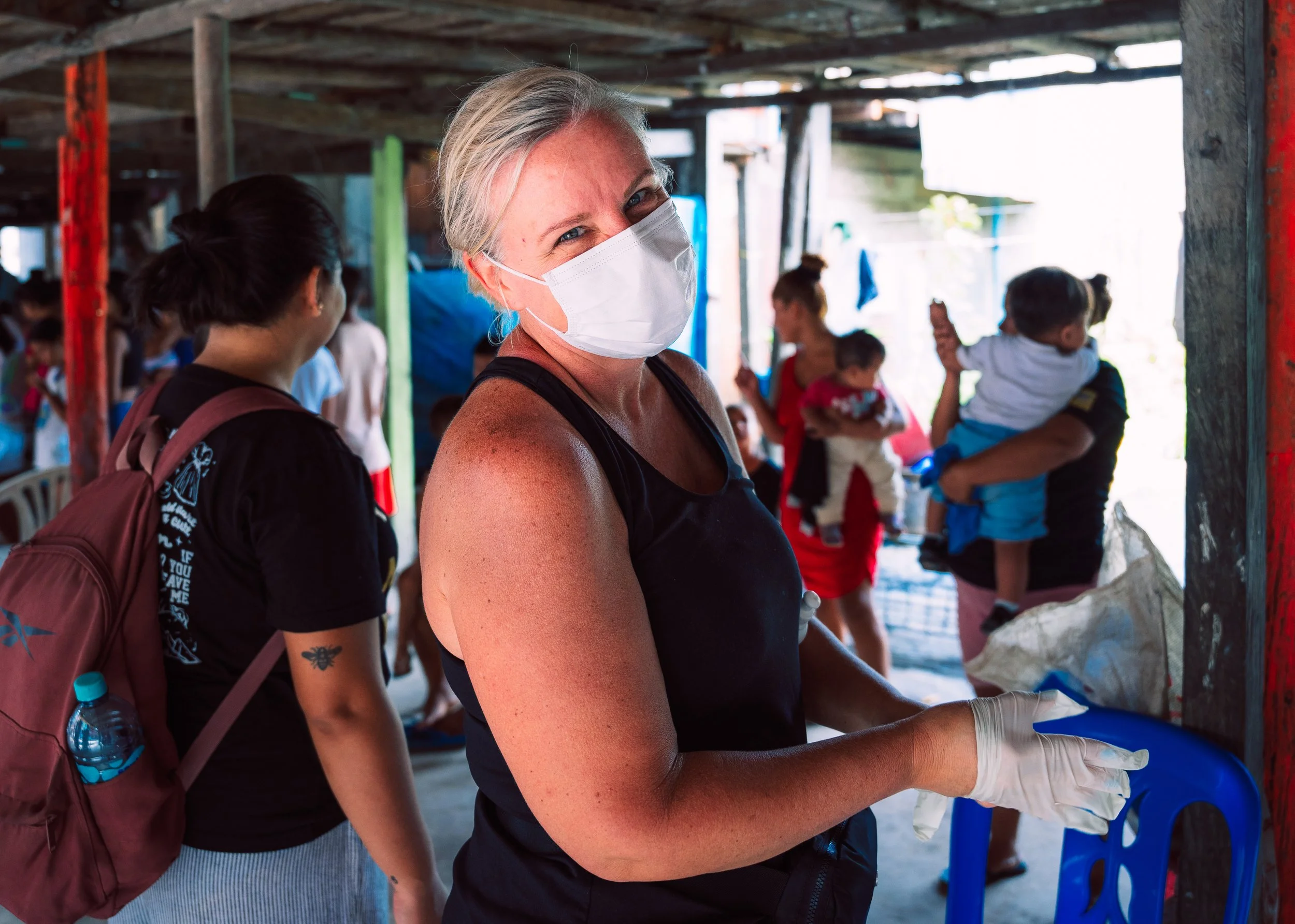 A woman wearing a white face mask and gloves, smiling at the camera, in a busy area with other people in the background, some carrying children and others wearing masks.