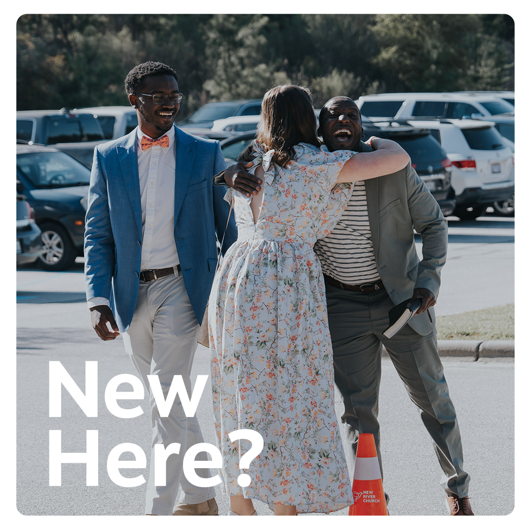 A woman in a floral dress hugging a man in a gray suit, smiling. A man in a blue suit with a bowtie and glasses is walking nearby, smiling. They are outdoors in a parking lot with cars in the background.