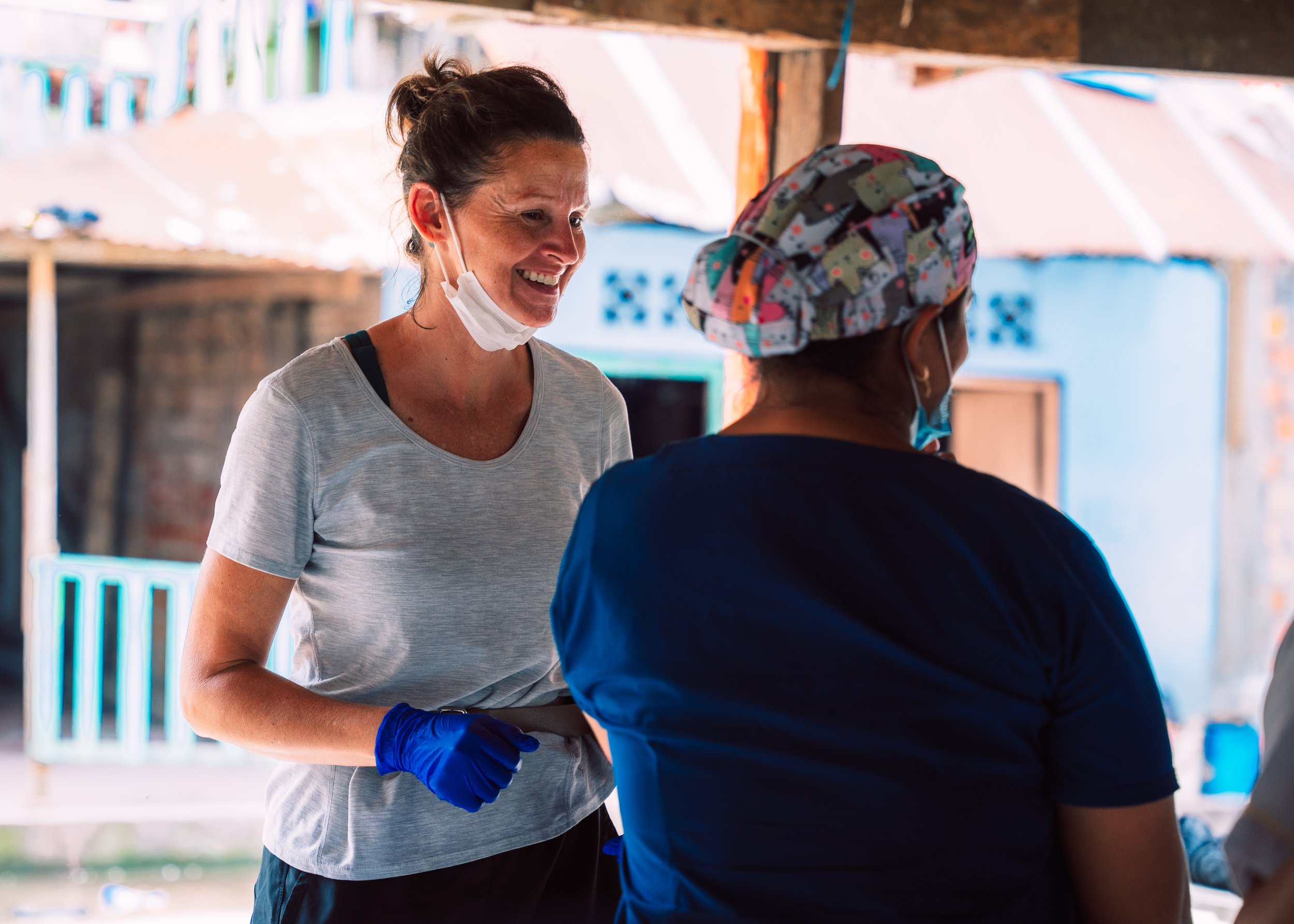 A woman smiling and talking to another woman wearing a colorful head covering, both with face masks, in an outdoor setting.