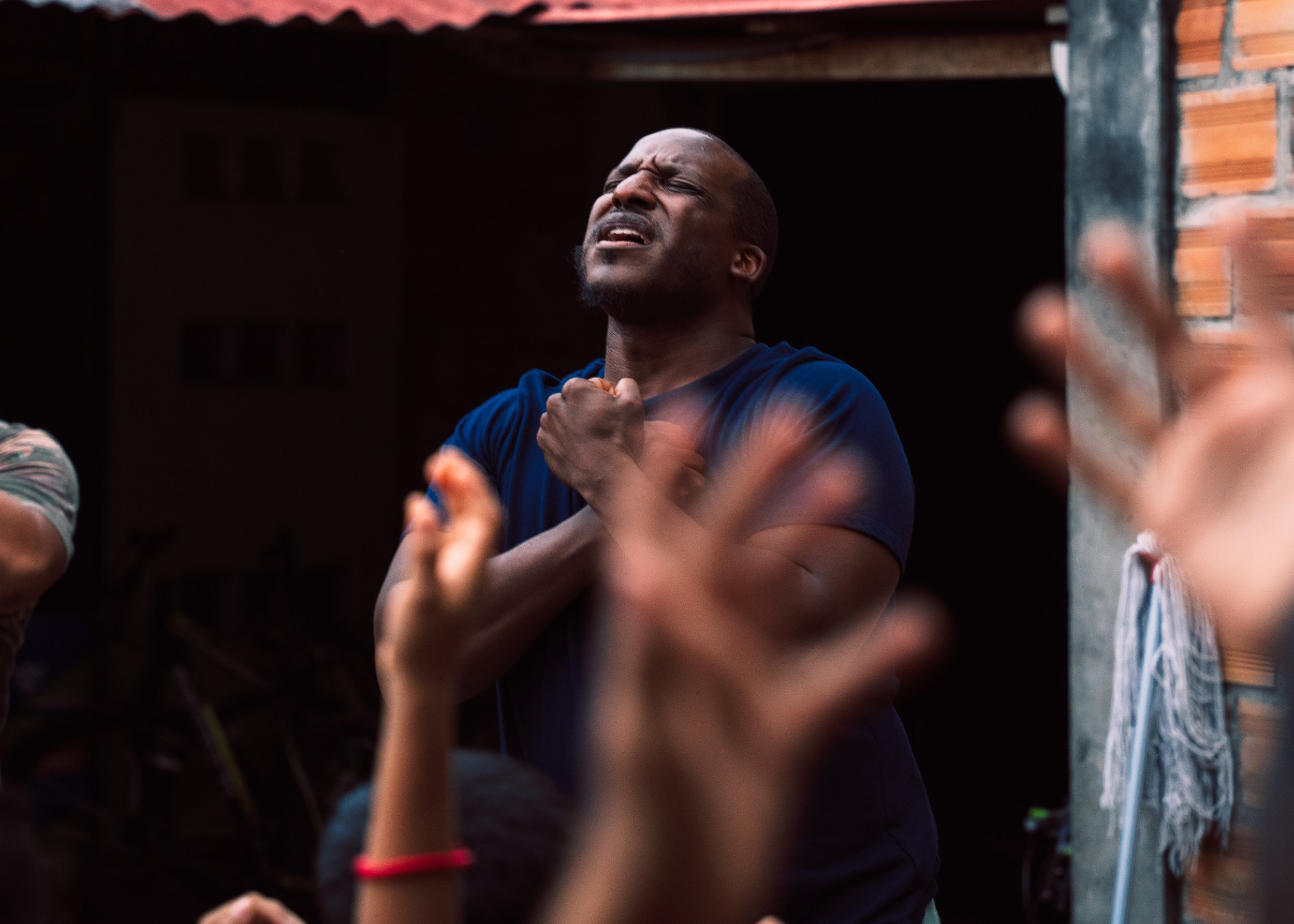 A man with closed eyes and a pained expression, holding his chest with one hand, appears to be experiencing discomfort or emotion during a gathering, with blurred hands and arms of other people in the foreground.