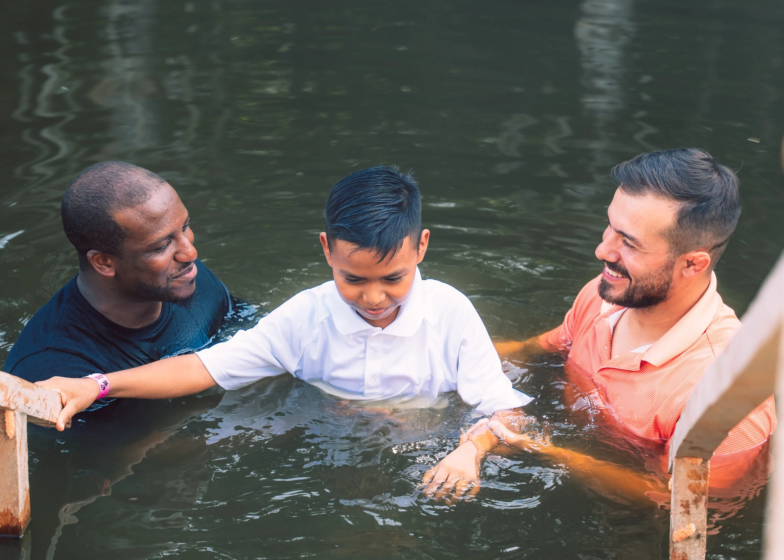 Three people, two men and a boy, are in a body of water, likely a river or pond, engaged in a religious baptism ceremony. The boy is being baptized by the two men, one on each side, with the man on the right holding him securely while the boy appears