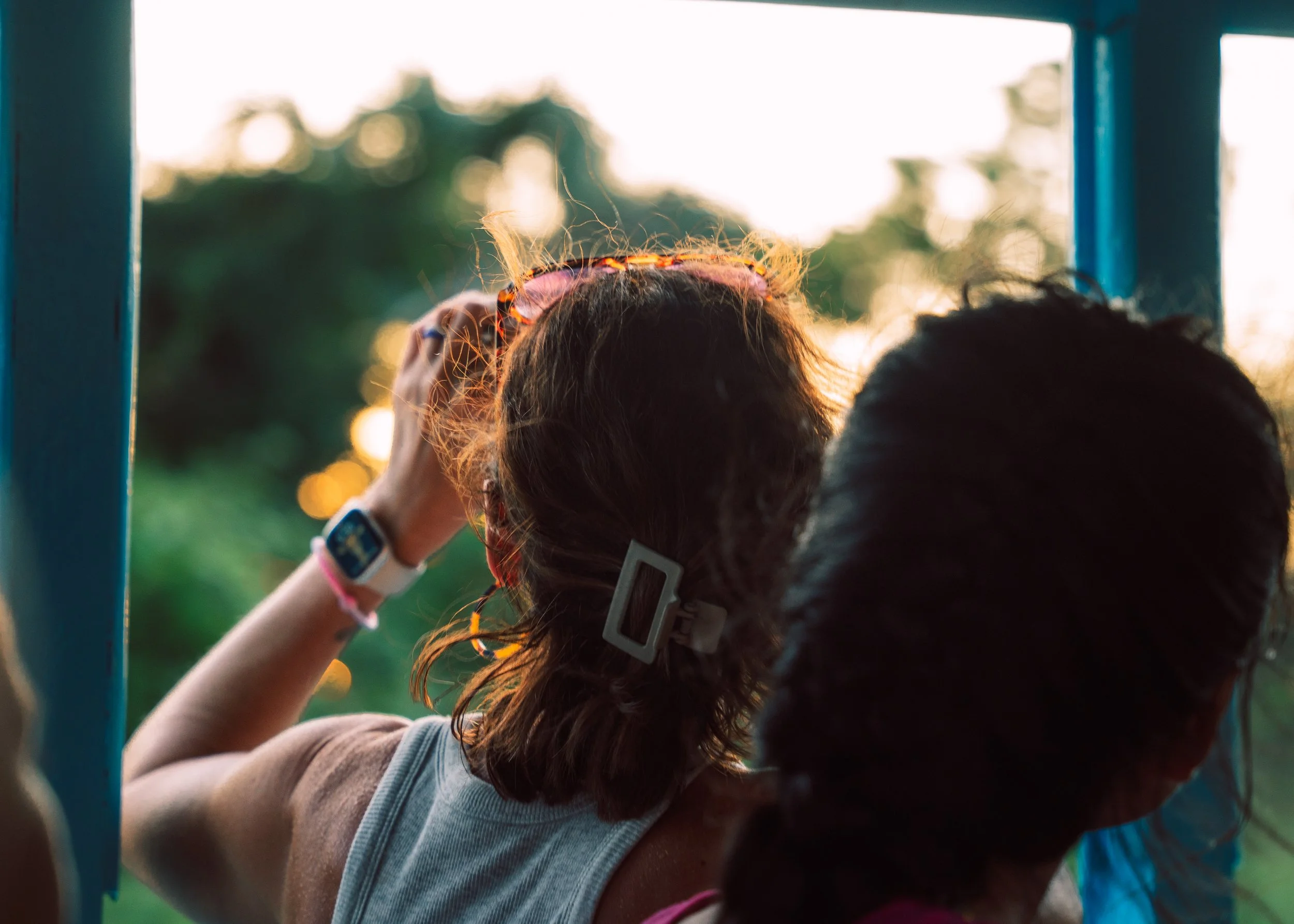Two young girls are looking outdoors through a blue metal frame, with sunlight and greenery in the background. One girl has shoulder-length hair with a clip and is holding sunglasses, while the other girl has long hair tied back.