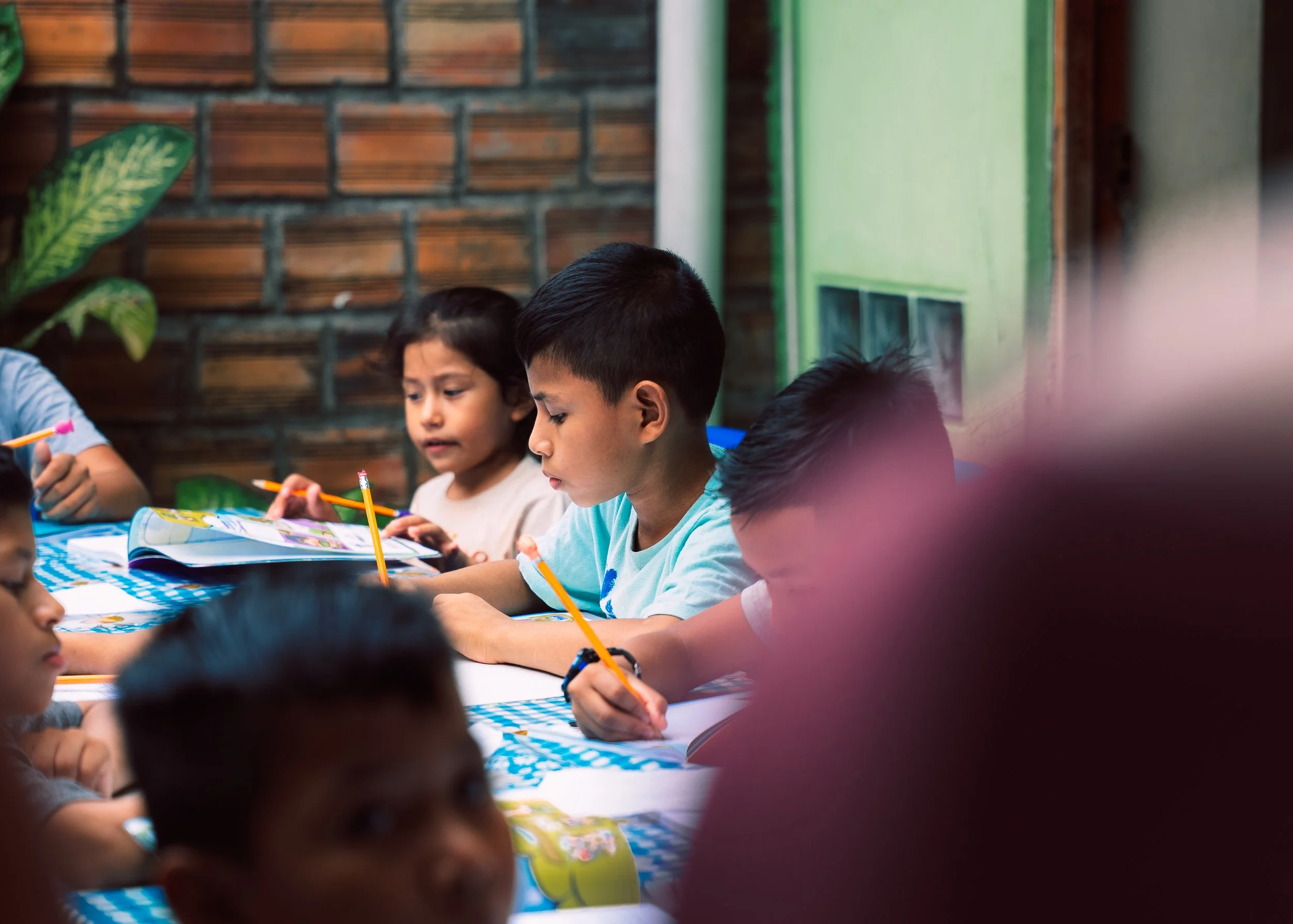 Children sitting at a table, engaged in reading or writing activities, with a brick wall and green door in the background.