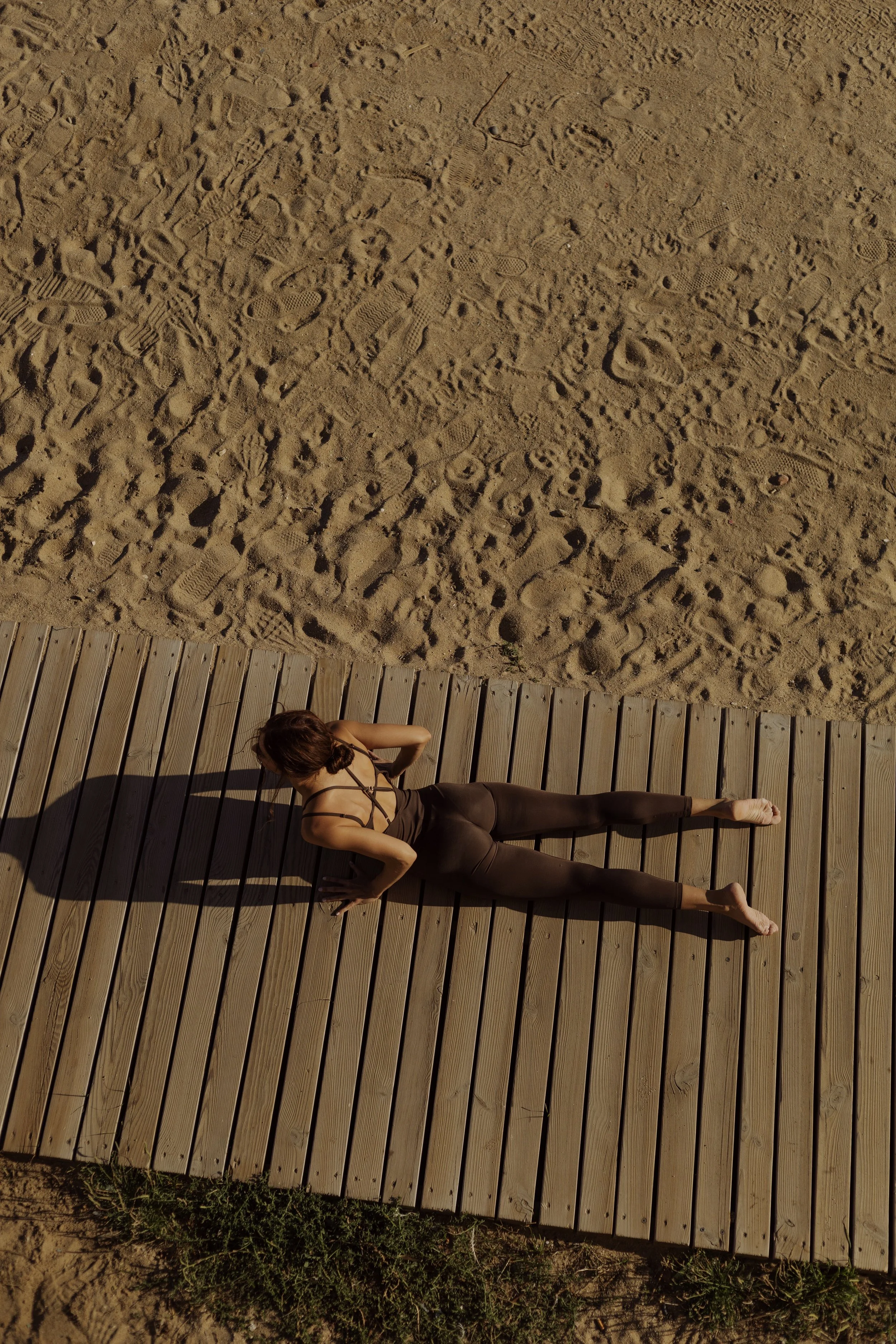 A woman practicing yoga outdoors on a wooden deck near a sandy area with numerous footprints, performing a side plank pose.