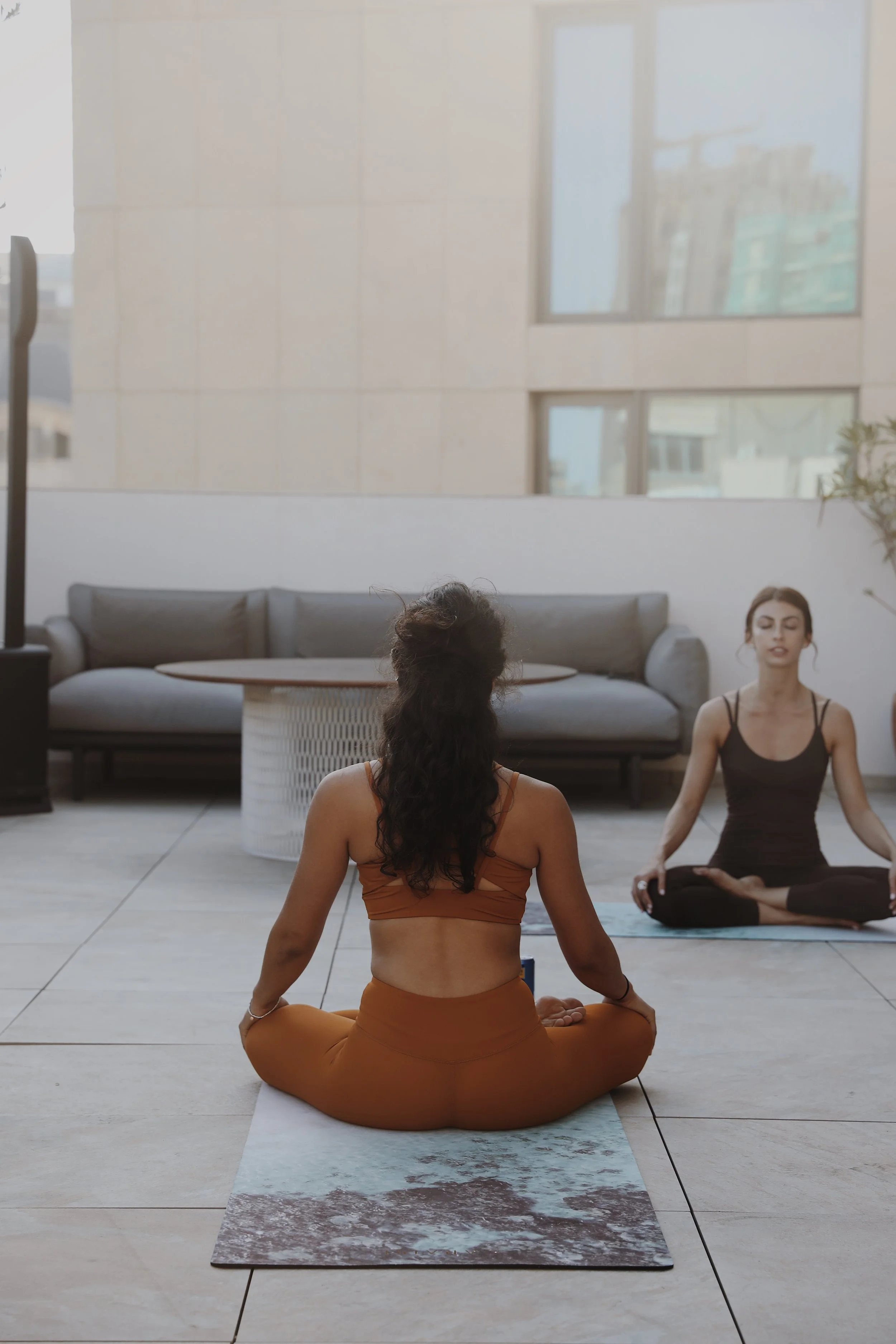 Three women practicing yoga outdoors on mats, sitting cross-legged with their eyes closed, in a peaceful urban setting with a modern building in the background.