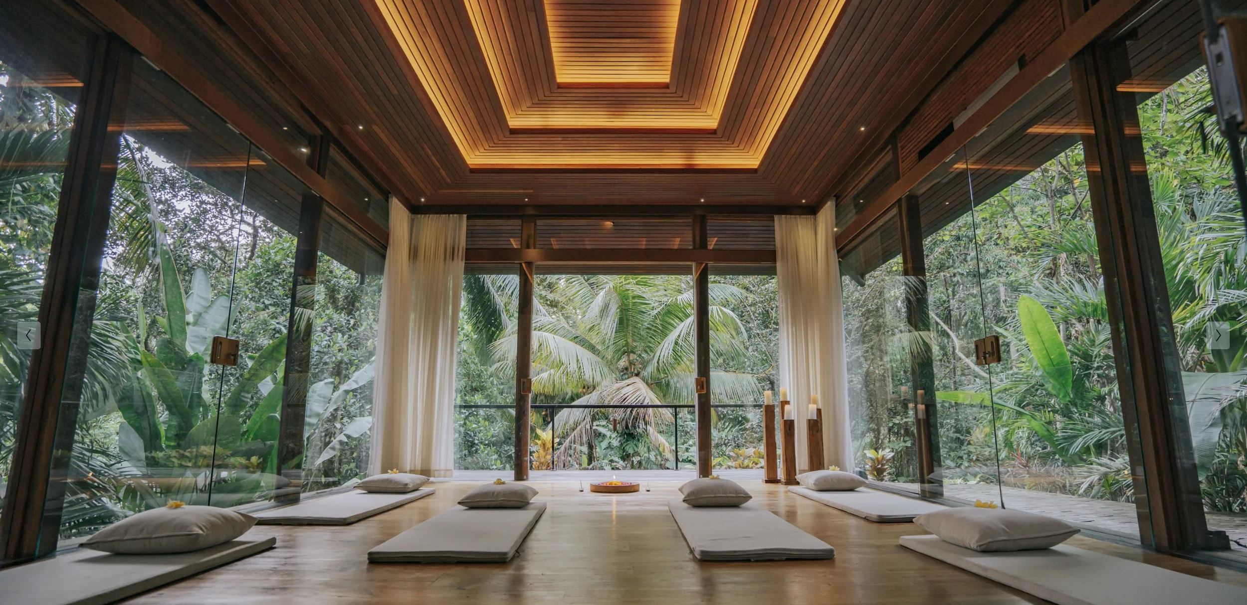 Indoor space with large glass windows, view of lush tropical greenery, wooden ceiling, and cushions on the floor for meditation.