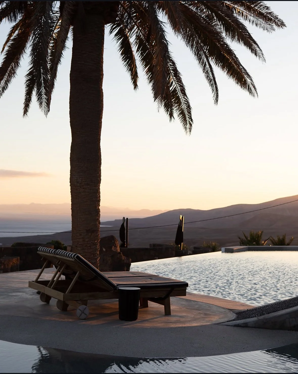 A scenic view of a sunset over a body of water with mountains in the distance, featuring a poolside scene with a palm tree, a lounge chair, and a small table.