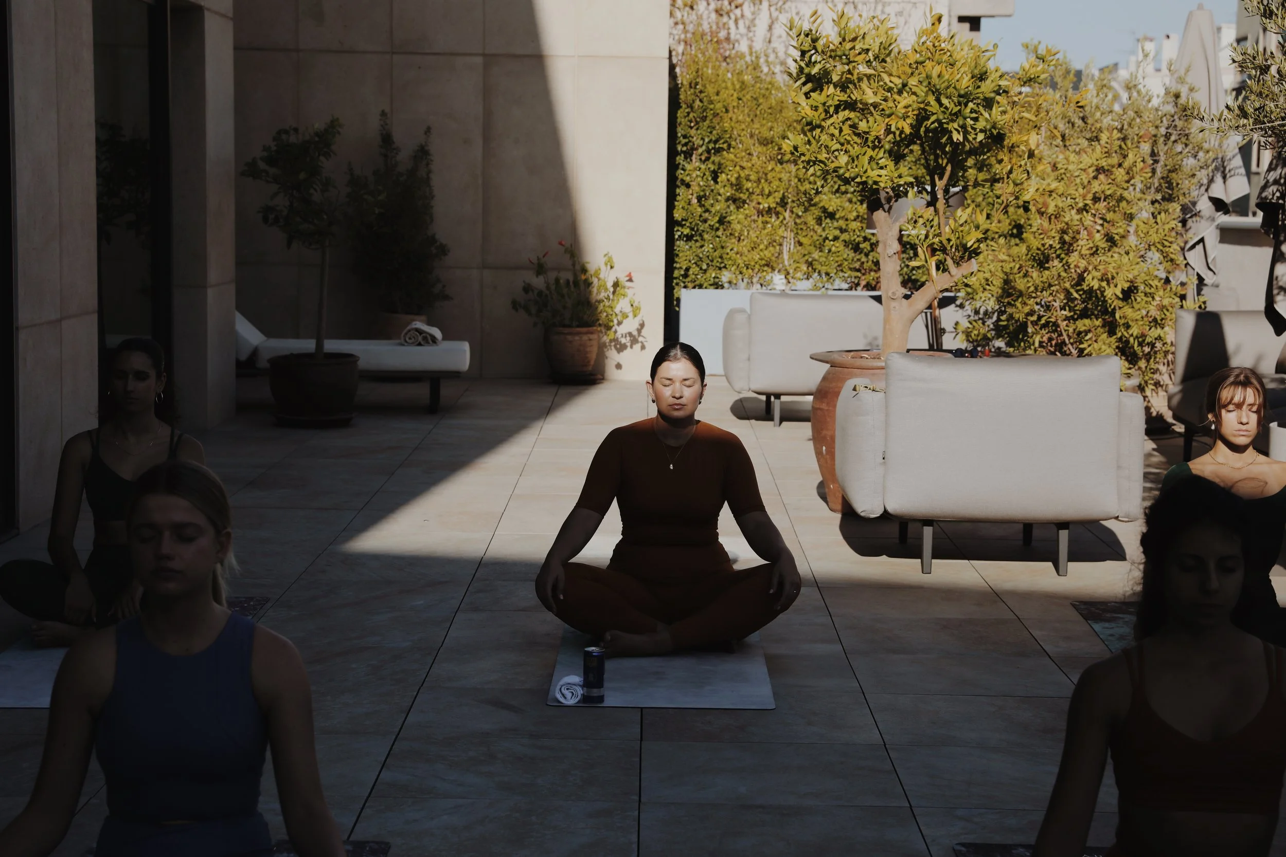 A group of people practicing meditation outdoors on a sunny day, seated cross-legged with eyes closed on a terrace surrounded by potted plants and trees.