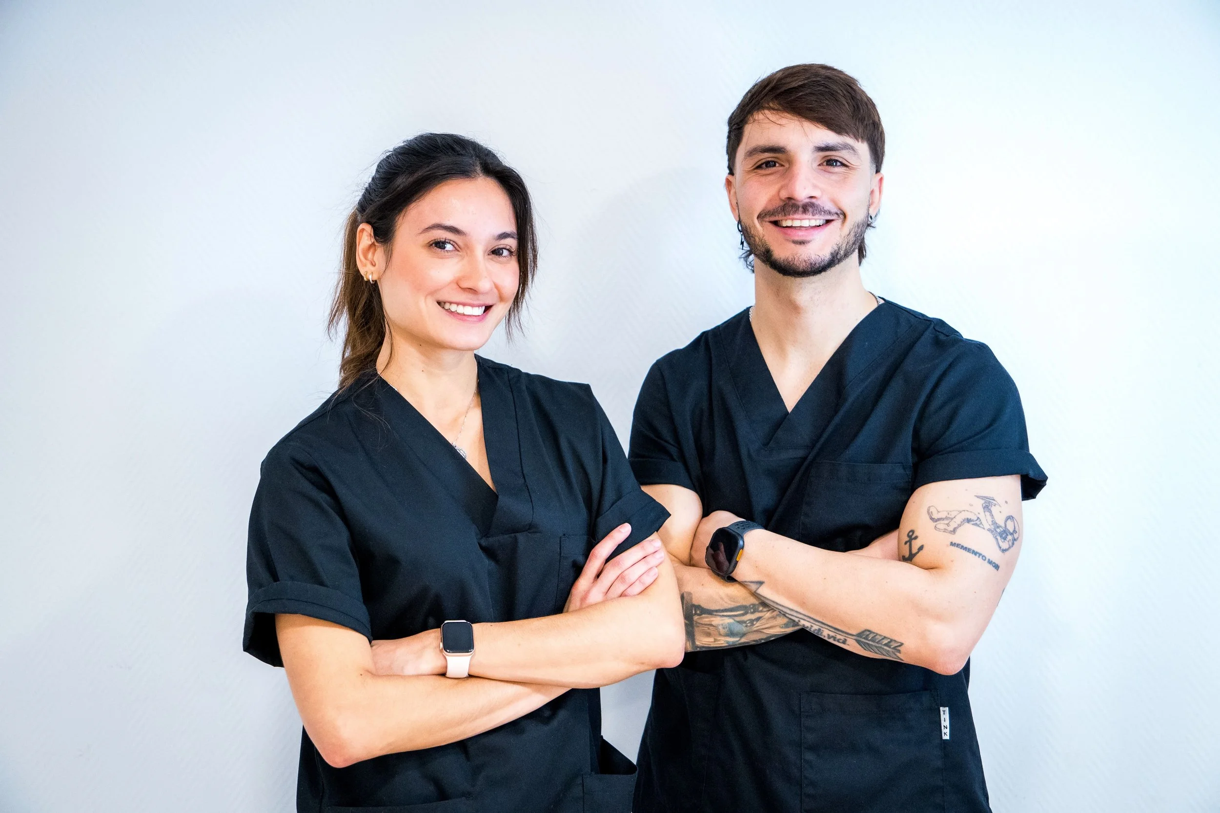Dos profesionales, una mujer y un hombre, vestidos con uniforme negro, sonriéndo y posando de pie con los brazos cruzados, frente a una pared blanca.