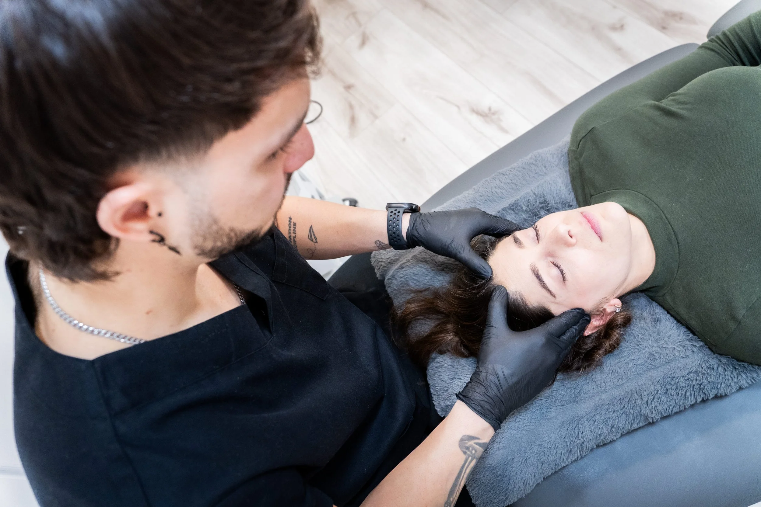 Un hombre que lleva guantes negros realiza un procedimiento estético en la cara de una mujer que está acostada, con los ojos cerrados y una toalla gris debajo de la cabeza.