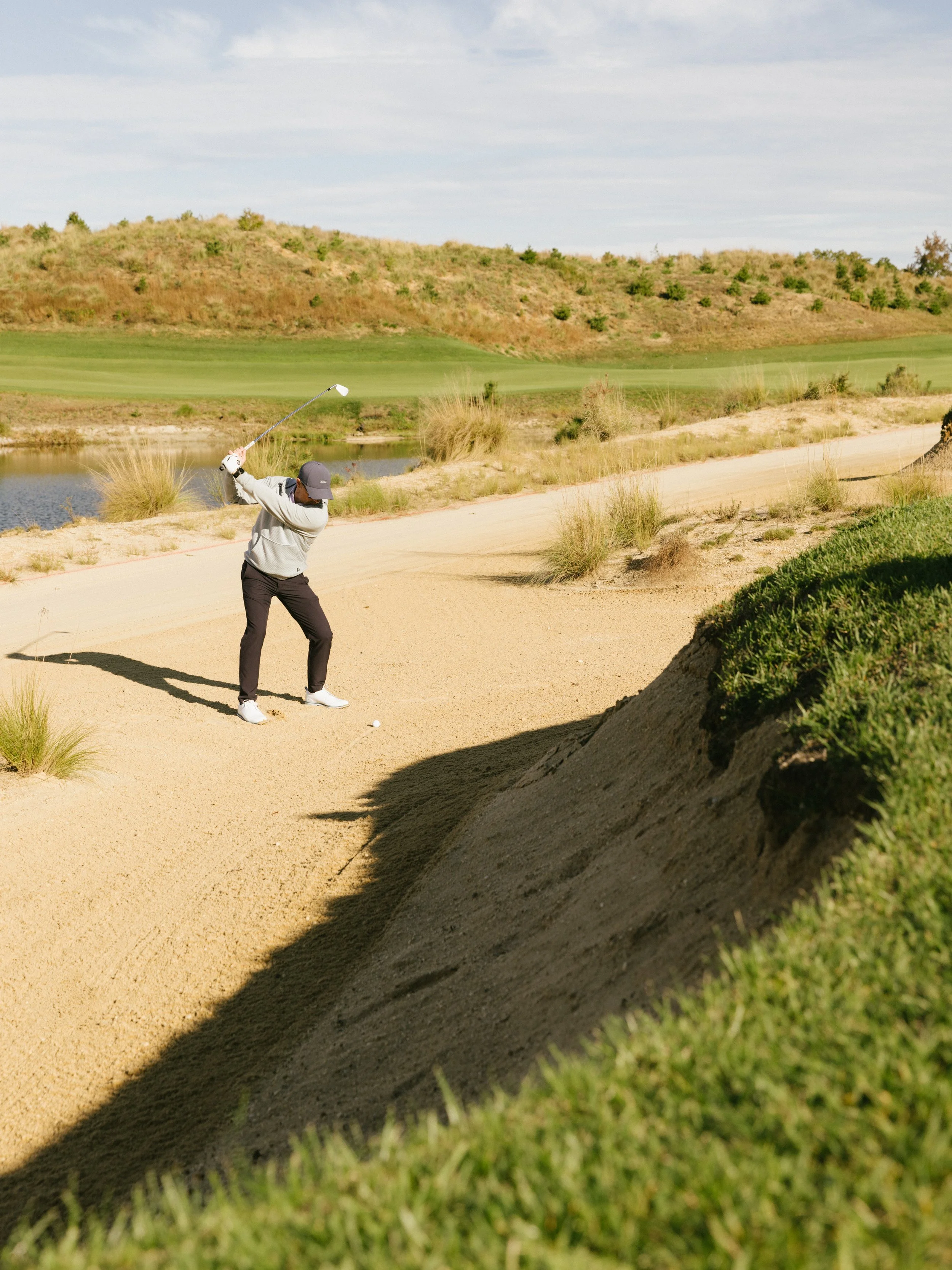 A man playing golf on a course near a sand bunker with grass patches and a water hazard, surrounded by hills with trees and a cloudy sky.