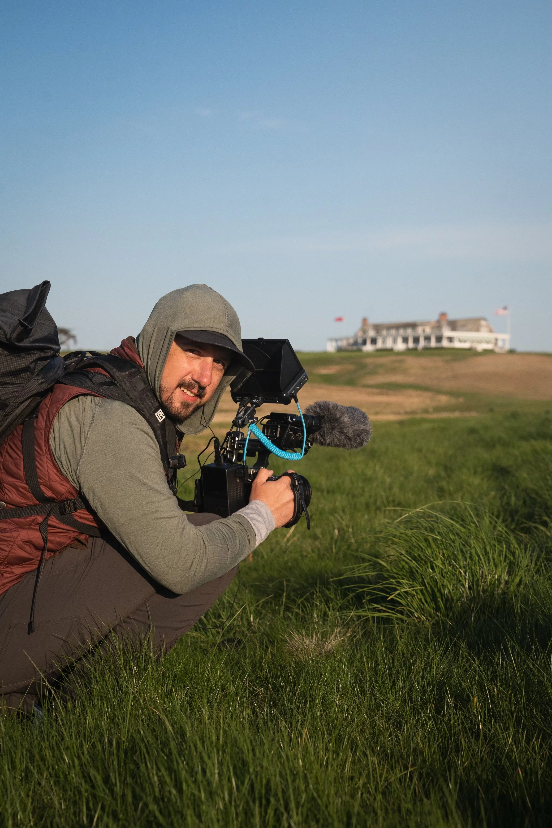 A man crouching in tall grass with a camera and microphone, with a large white house on a hill and flags in the background.