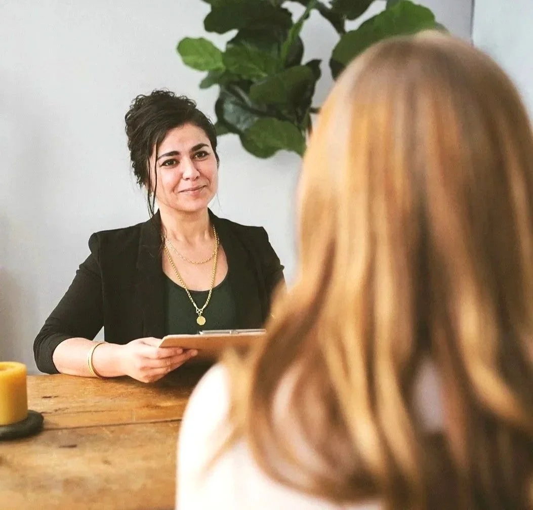 A woman with dark hair and a black blazer is sitting at a table, holding a tablet, and looking at another woman with long, light brown hair. There is a green plant in the background and candles on the table.
