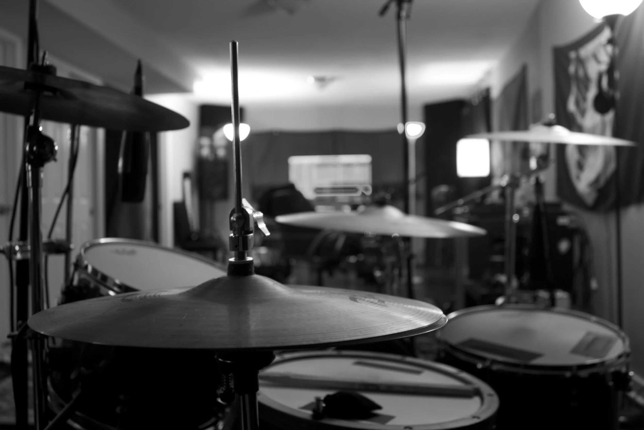 A recording studio with a drum set in the foreground, hi hats in focus, with a mixing desk, speakers, and guitar amp in the background