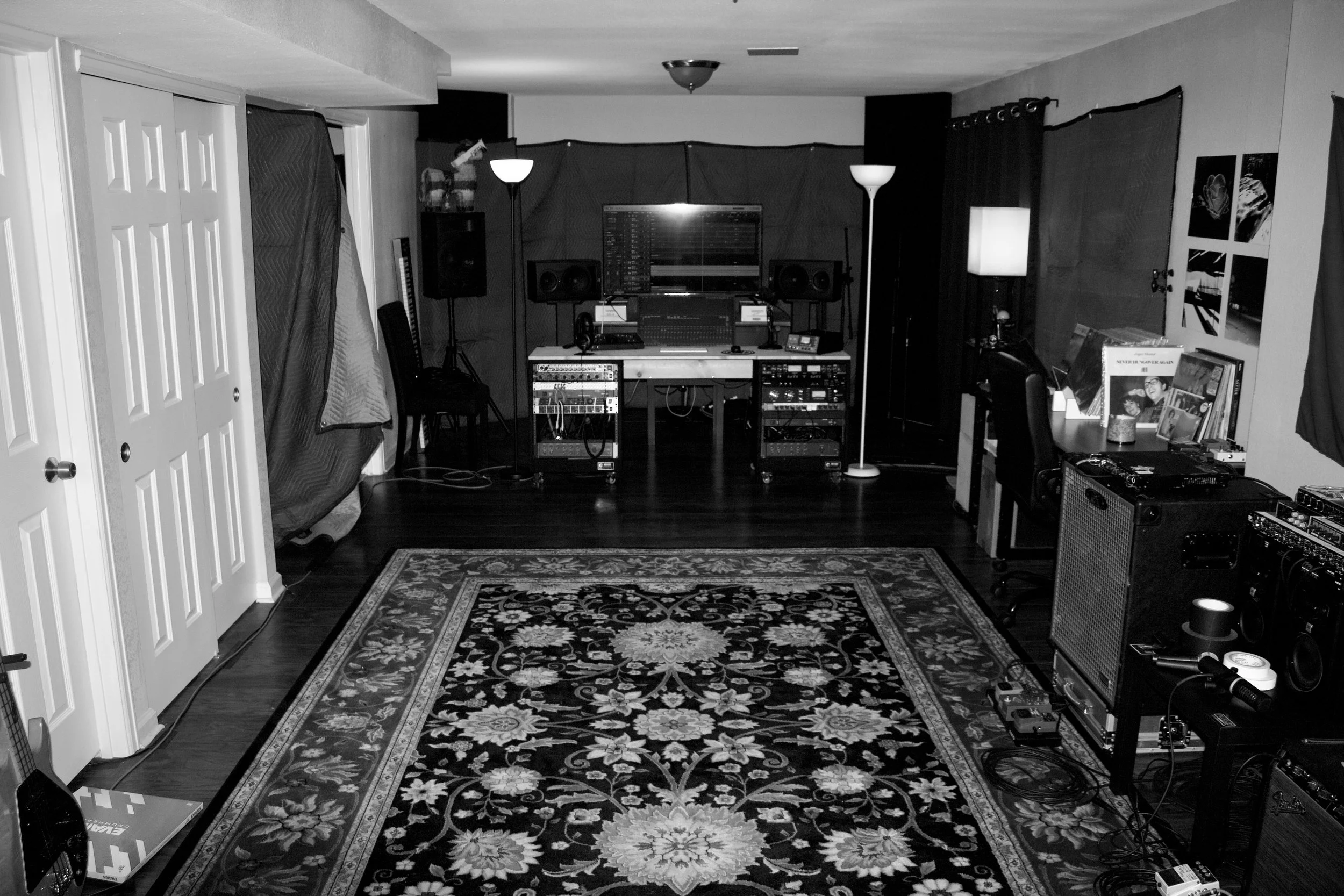 A Recording studio with a rug, bass amplifier, vinyl records, mixing desk, studio gear, rack gear, lamps, computer monitor, and a bass guitar.