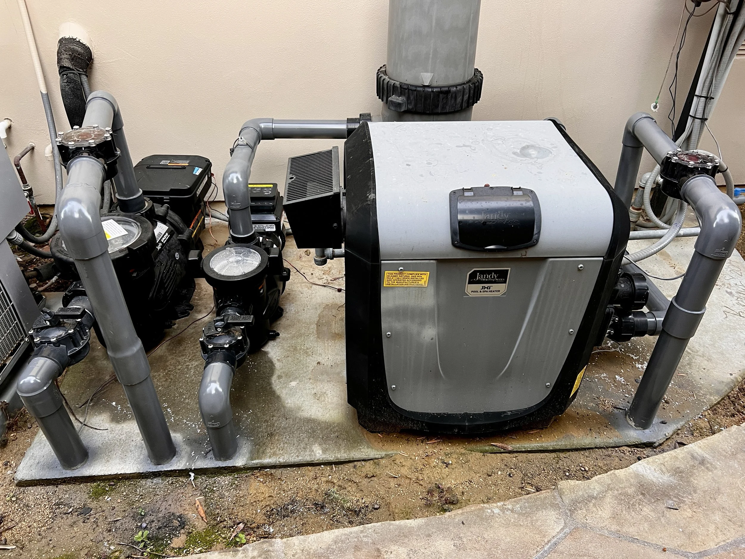 An outdoor swimming pool pump system with grey pipes and a large pool filter in a concrete pad.