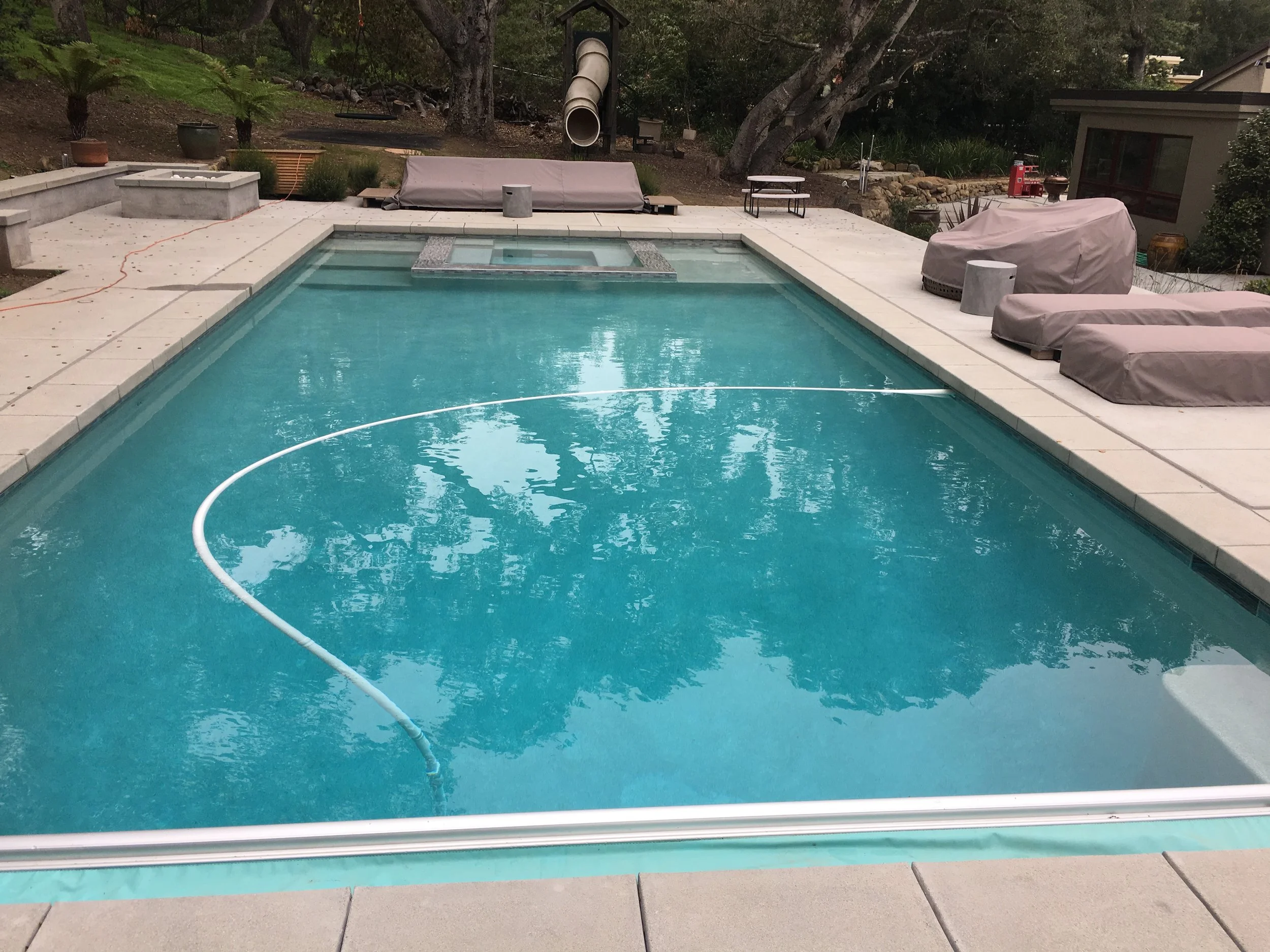 A backyard swimming pool with a white pool cleaning pole floating on the water. The pool has a raised hot tub at one end, and there are covered outdoor furniture and potted plants on the patio surrounding the pool. Trees and a house are visible in the background.