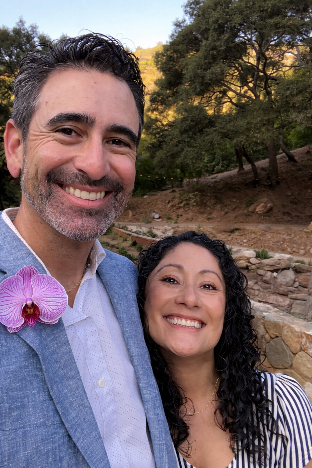 A smiling man with dark, curly hair and a beard stands next to a smiling woman with curly hair in an outdoor setting with trees and a stone wall in the background.