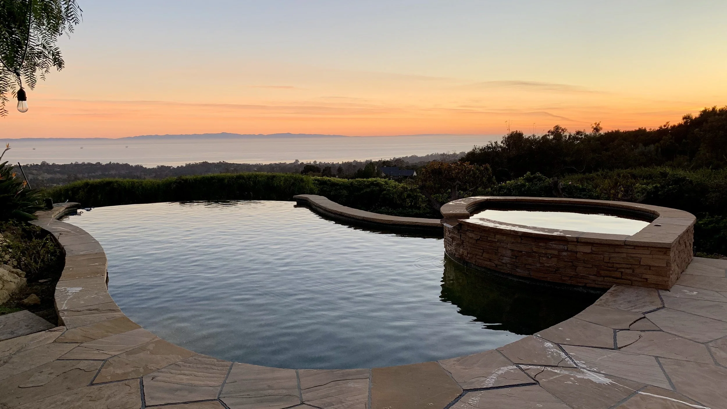 An infinity pool with a stone border overlooking a scenic sunset view of the ocean, with trees and hills in the background.