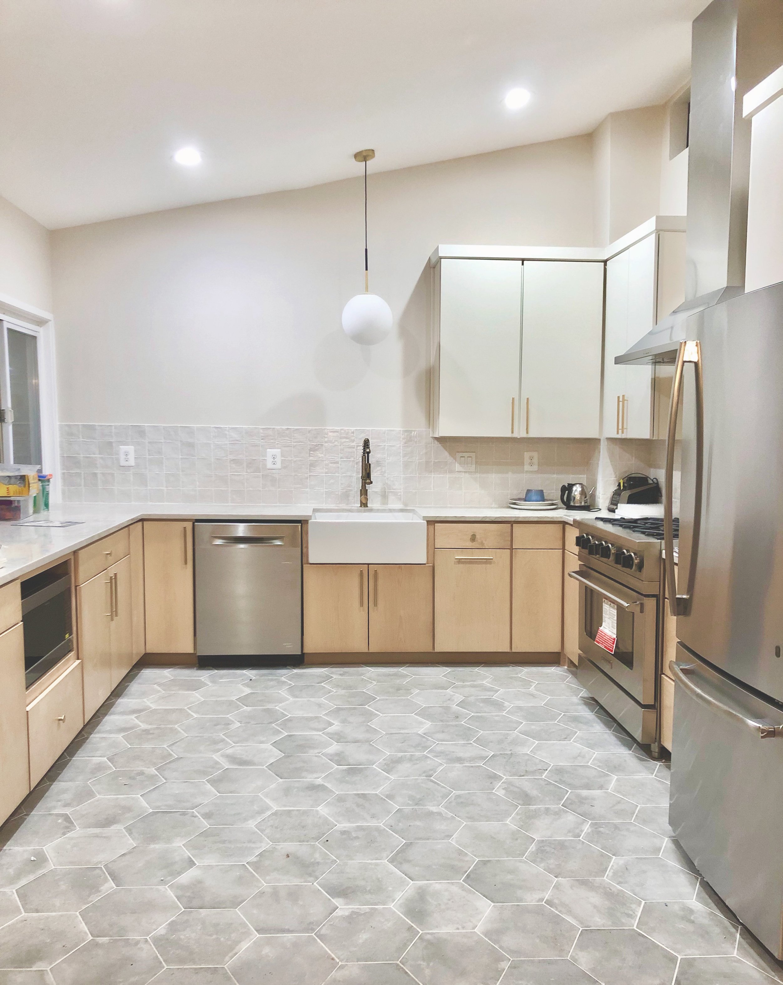Bright, modern U-shaped kitchen with raised ceiling, hex tile floor, light wood cabinets, and a central farmhouse sink.