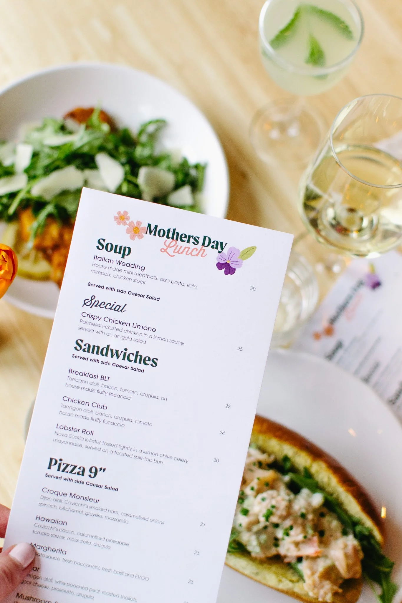 A person's hand holding a fork over a mini cheese quiche on a white plate, garnished with mixed greens, with pink flowers in the foreground.