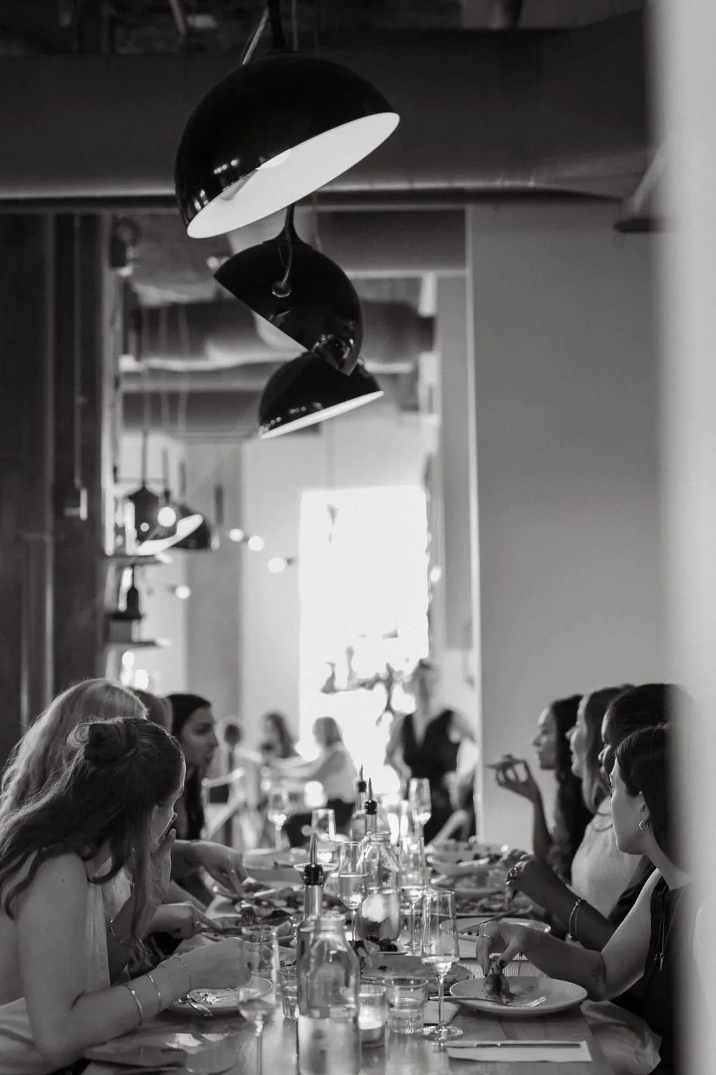 Interior of a busy restaurant with booths, hanging pendant lights, and patrons dining, some engaged in conversation, with large windows and decorative lighting.