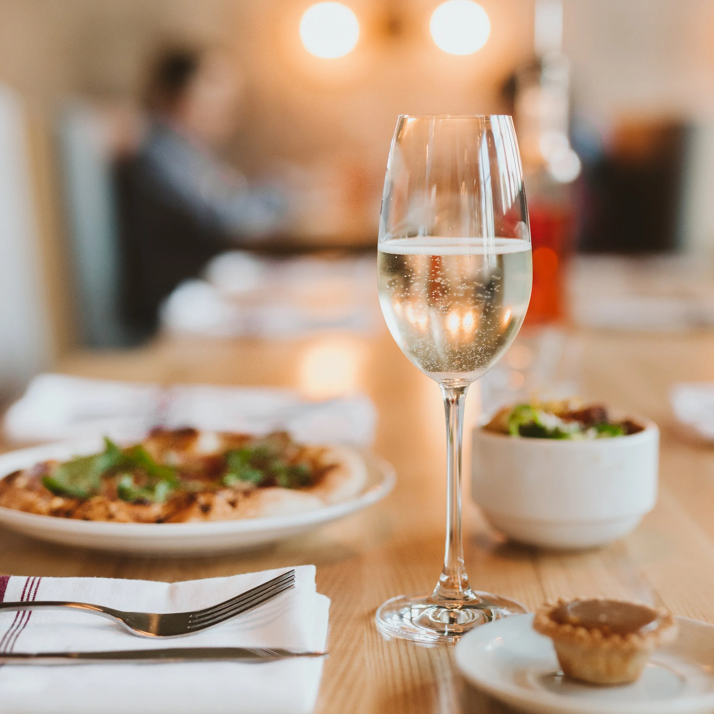 A person's hand holding a fork over a mini cheese quiche on a white plate, garnished with mixed greens, with pink flowers in the foreground.