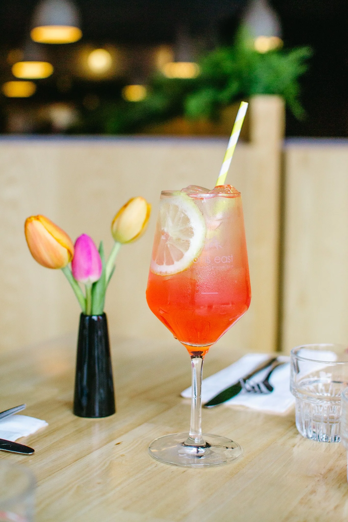 A woman holding a lemon slice in a glass of lemonade with a yellow straw. There is a pink cocktail and a can of lemon soda on the table.