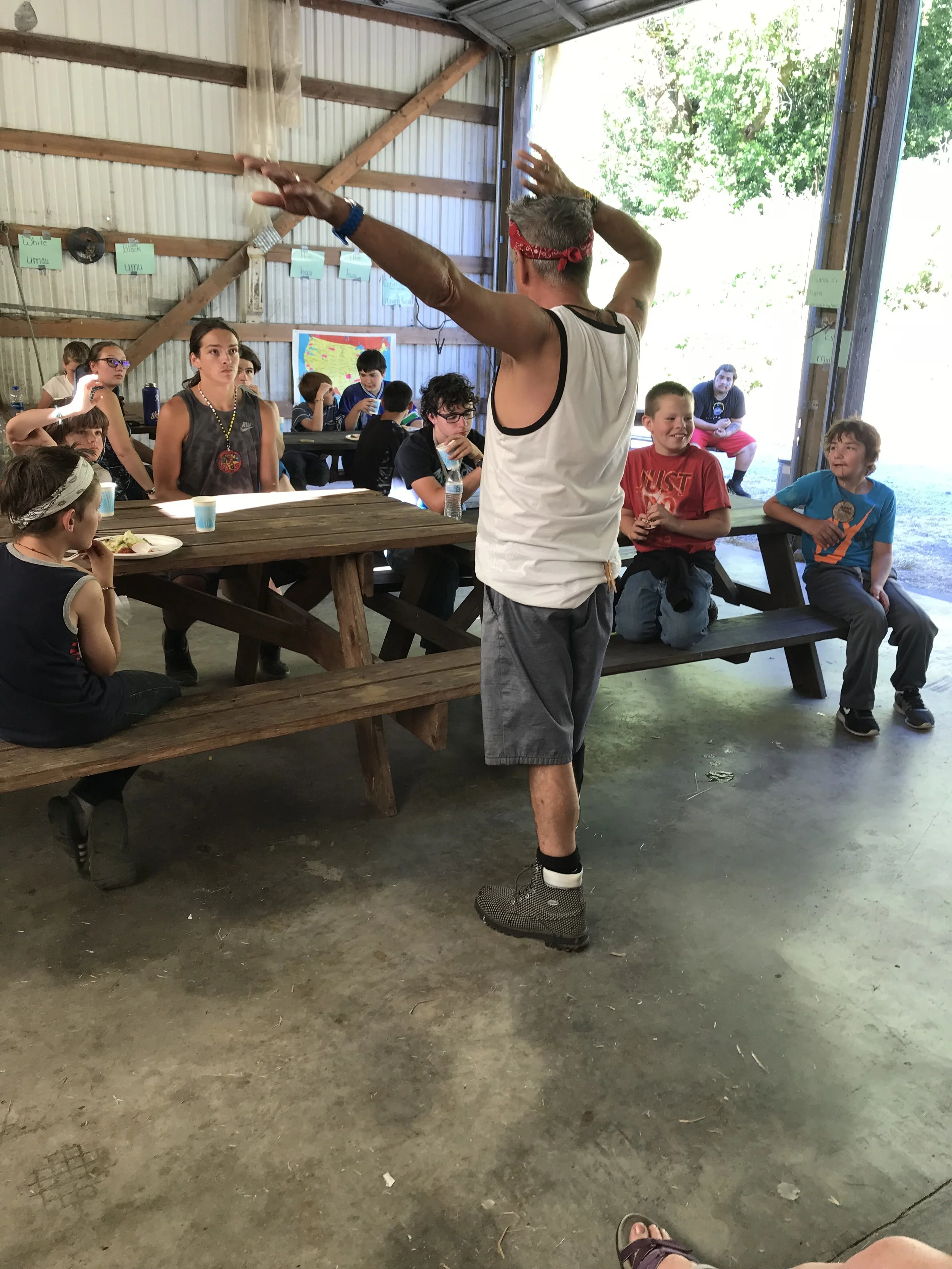 A man wearing a tank top, shorts, and a red bandana conducts a group activity with children inside a rustic barn or pavilion with open sides, wooden beams, and a dirt floor.