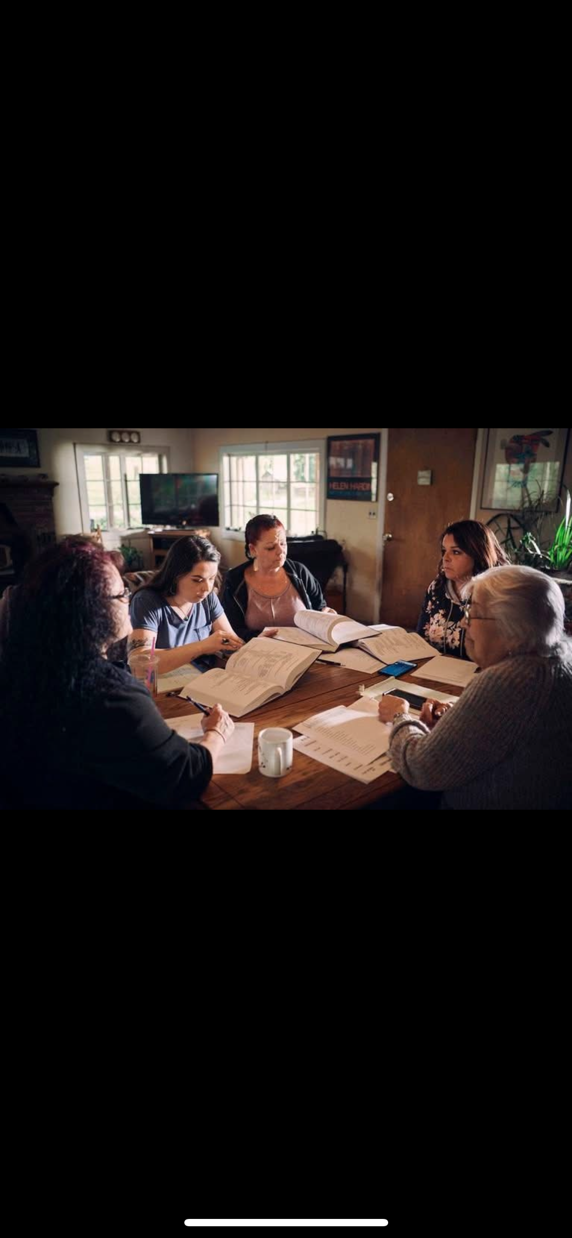 Five women sitting around a wooden table in a well-lit room, studying and reviewing large open books and papers.