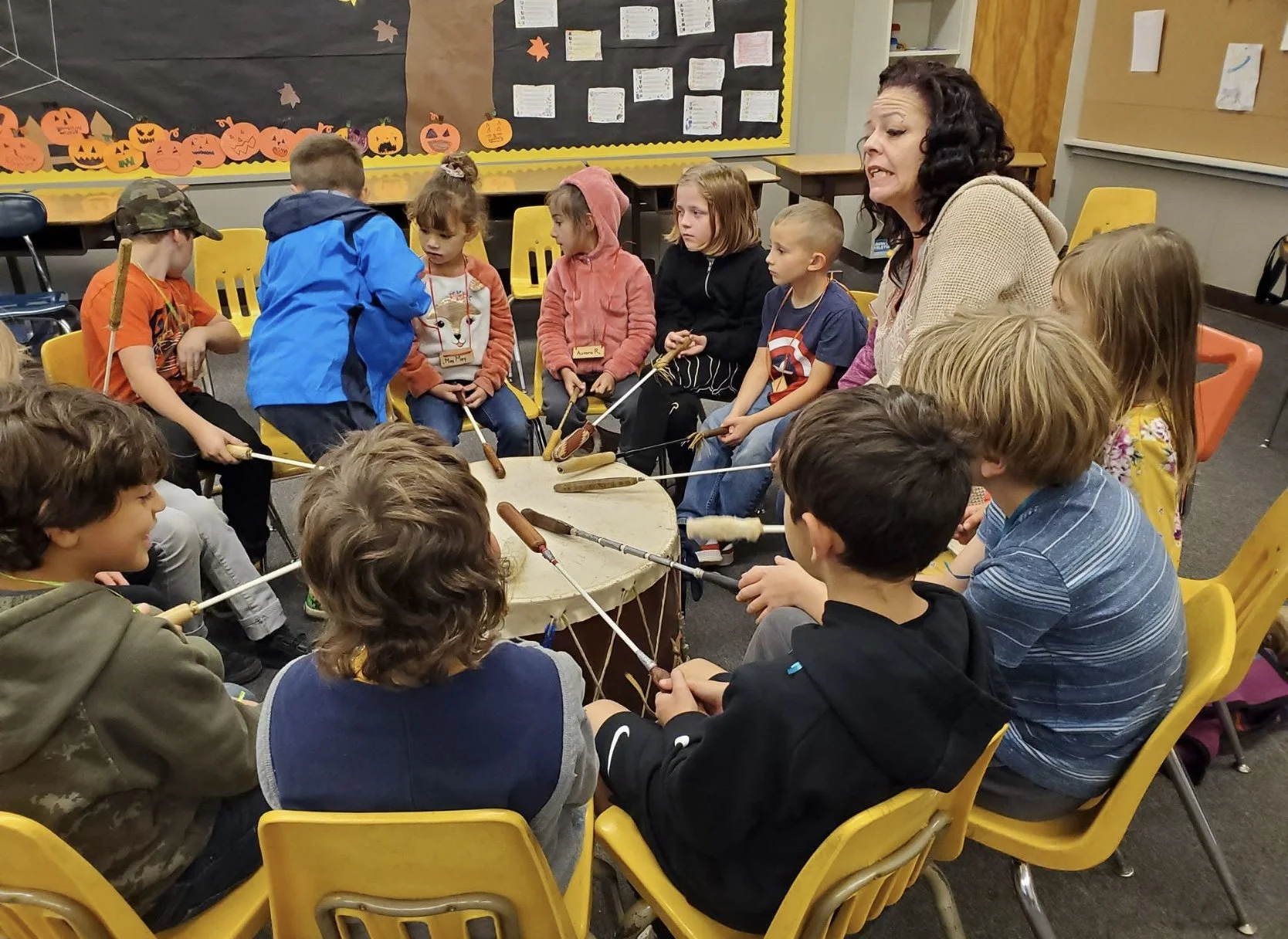 A group of children and a woman sit around a large drum, each holding a drumstick, participating in a drum circle activity in a classroom decorated with Halloween-themed artwork.