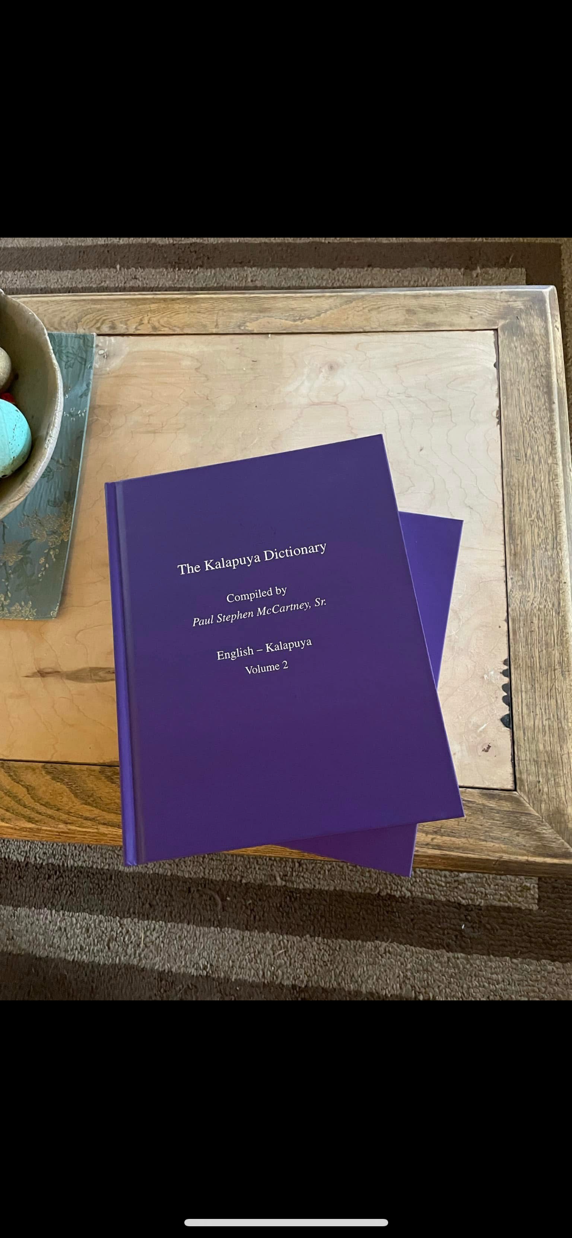 A purple book titled 'The Kalapuya Dictionary' on a wooden table, with part of a bowl of eggs visible on the left.