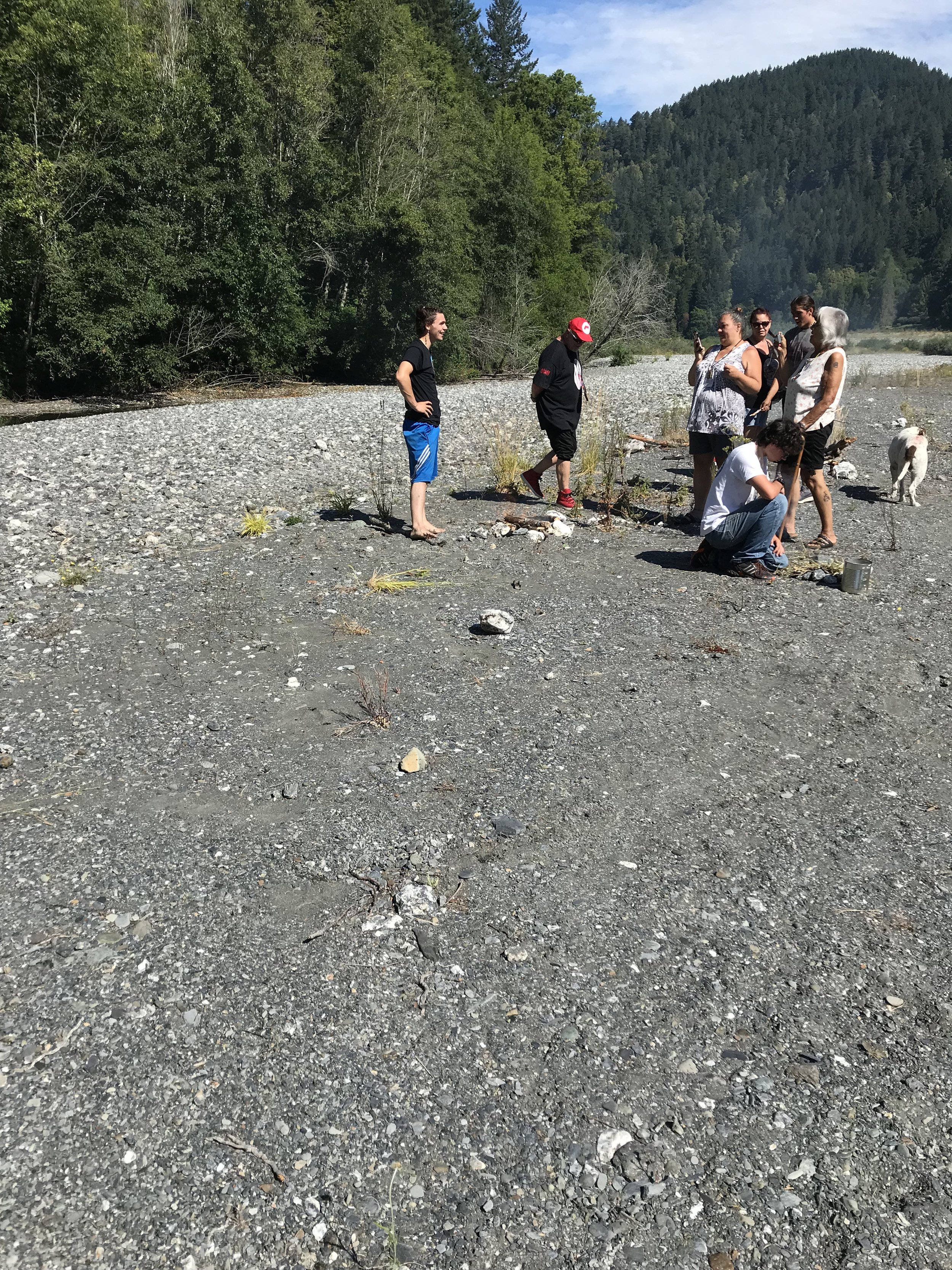 A group of seven people on a rocky riverbank surrounded by trees and mountains, some standing, some crouching, with two dogs nearby, the sky partly cloudy.