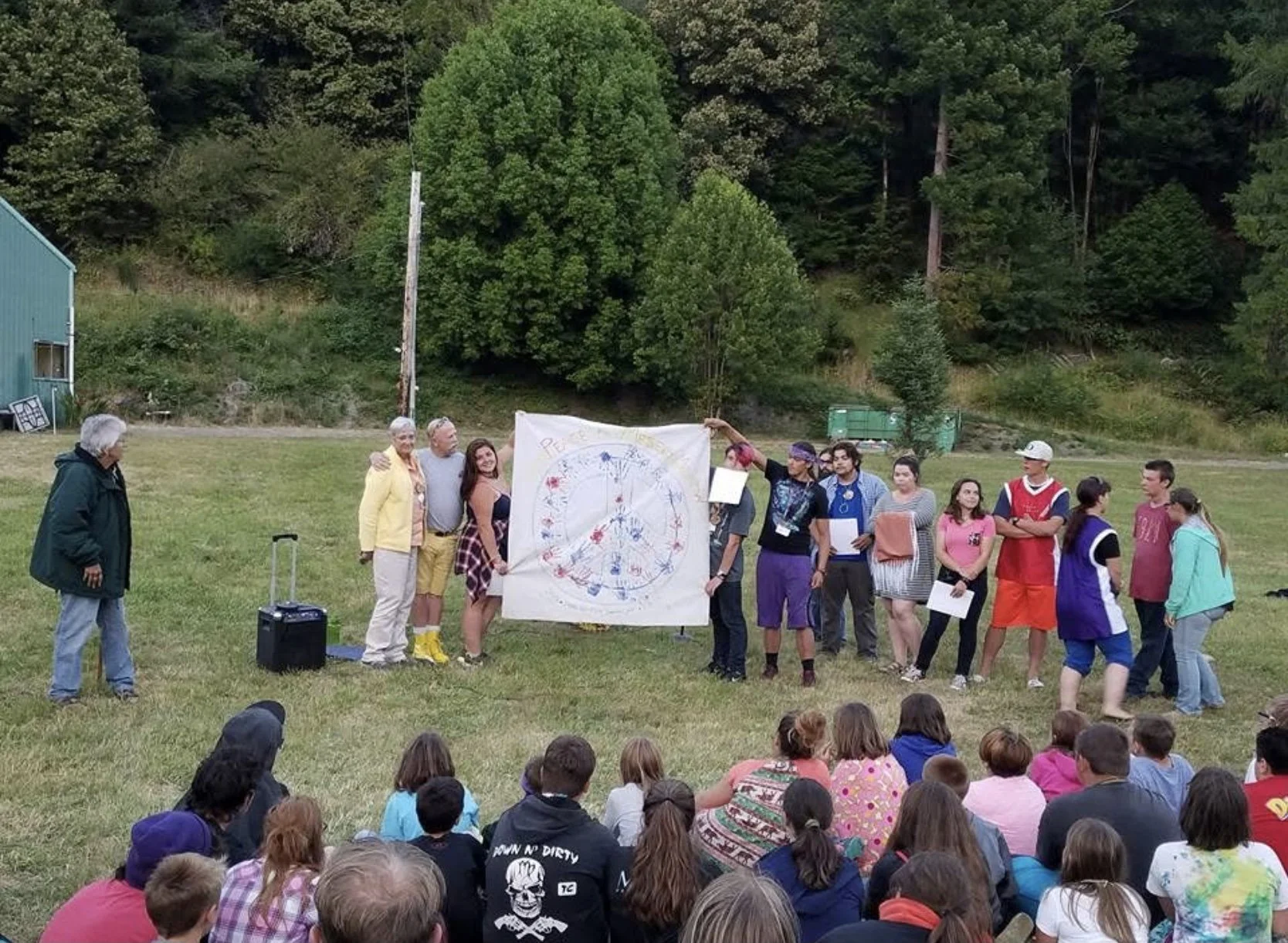 Group of people outdoors on a grassy field, holding a large circular artwork with various red, blue, and yellow markings, with children sitting on the grass in the foreground and trees in the background.