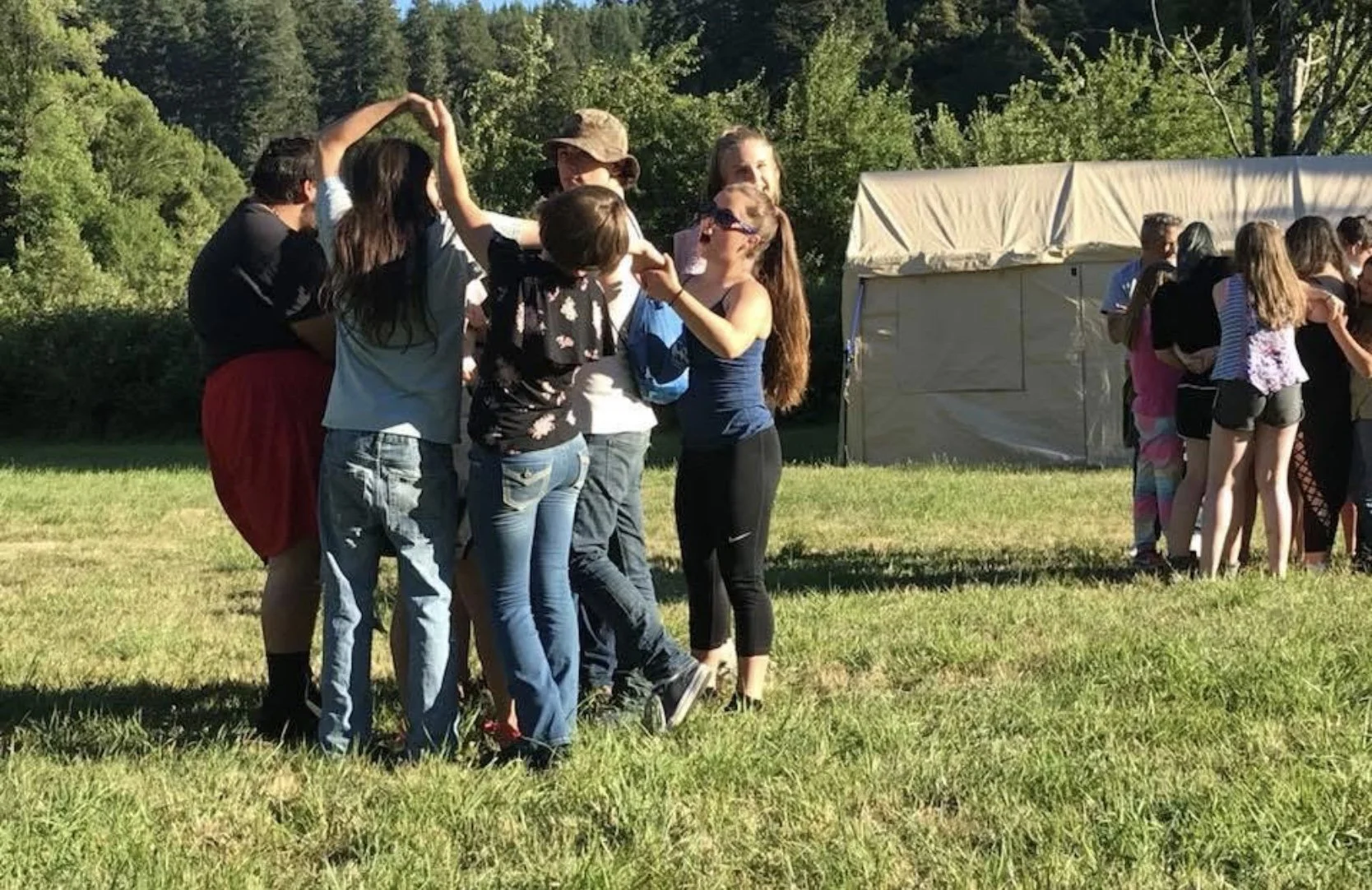 Group of people, mostly children, standing outdoors on grass in a line. Some are hugging or holding hands and appear to be in a casual, social setting. There is a tent in the background surrounded by trees.