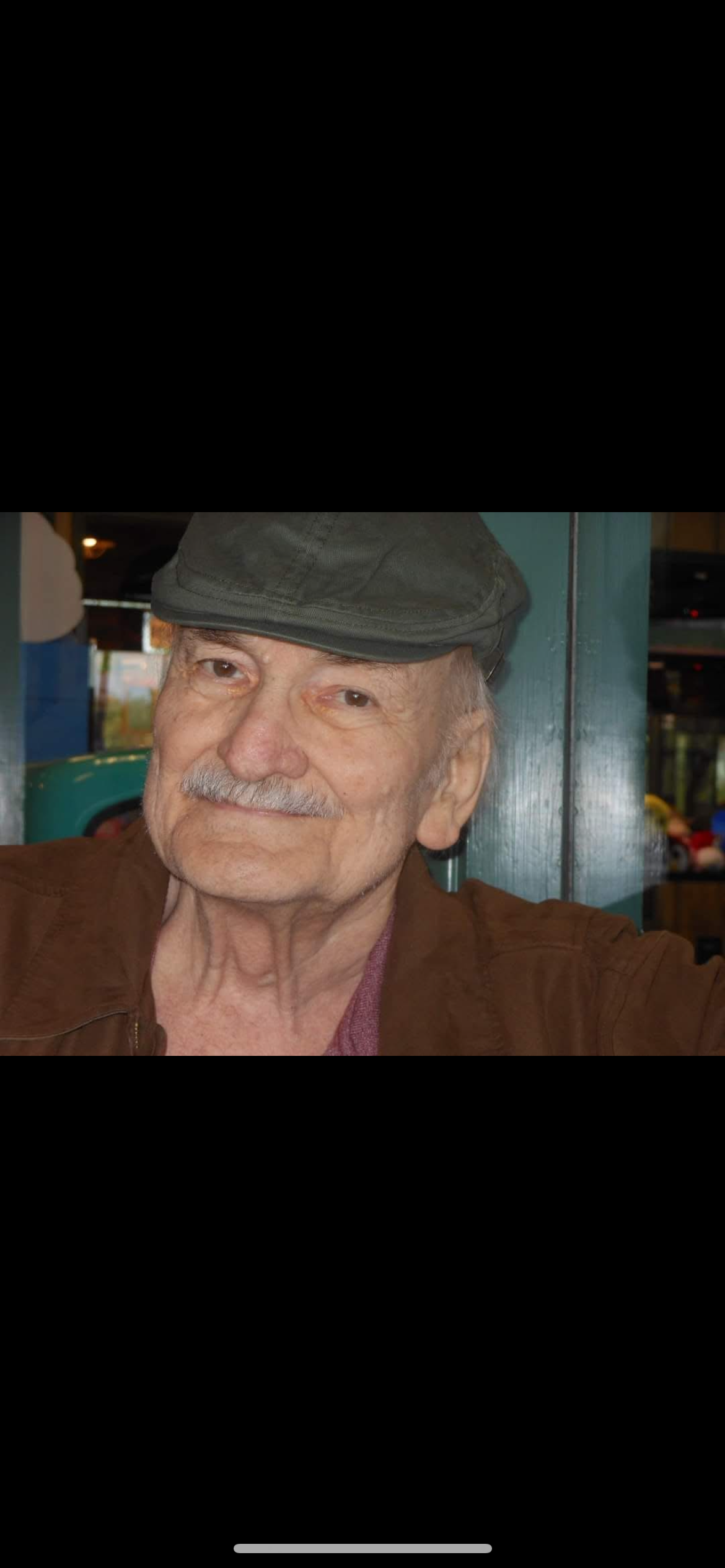 An elderly man wearing a dark green hat and a brown jacket, smiling softly, sitting indoors with a green structure and blurred objects in the background.