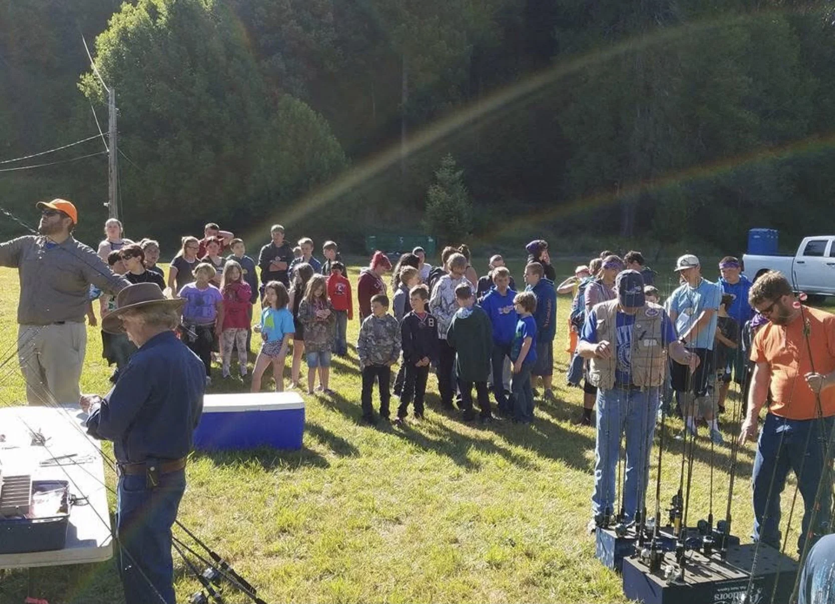 A large group of children and adults gathered outdoors on a grassy field, with some holding fishing poles, suggesting a fishing event or workshop. There are trees and a truck in the background, and the scene is illuminated by sunlight with rainbow-like lens flares.