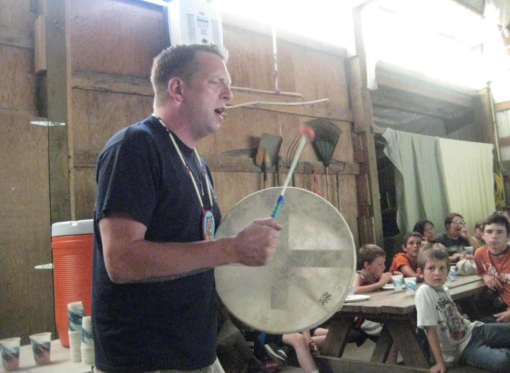 Man playing a large drum for a group of children in a rustic indoor setting.