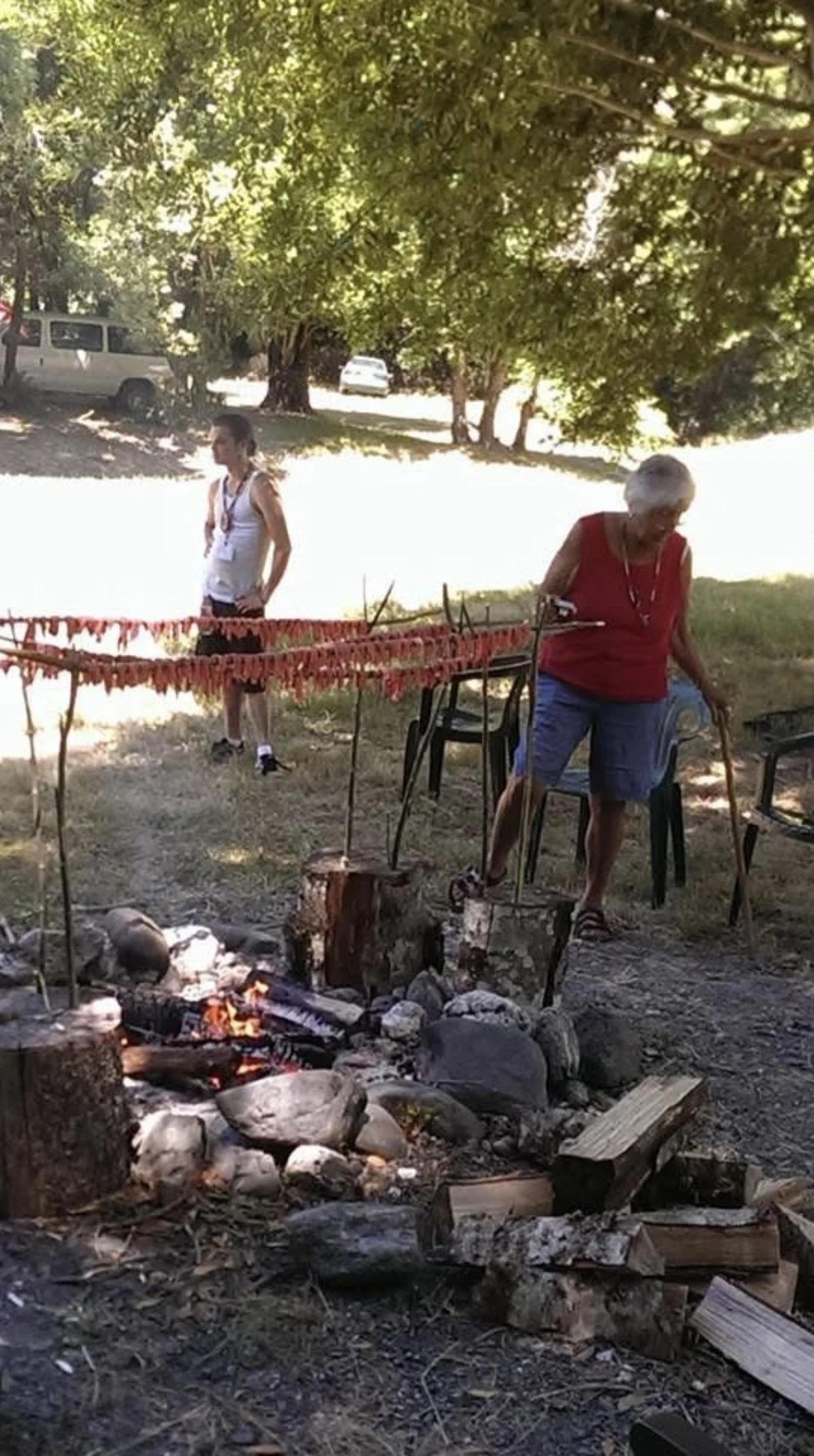 A woman with gray hair and a red sleeveless top stands near a campfire with rocks around it, outdoors under a large tree, while a younger woman in a white tank top stands in the background.