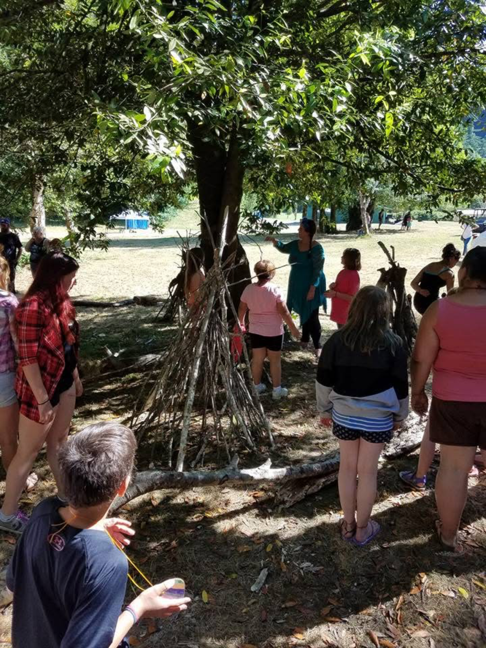 People gather around a large tree in a park, with some making a structure from branches and sticks. A woman appears to be explaining something to the group. The scene is outdoors in daytime, with several trees and a grassy area visible.