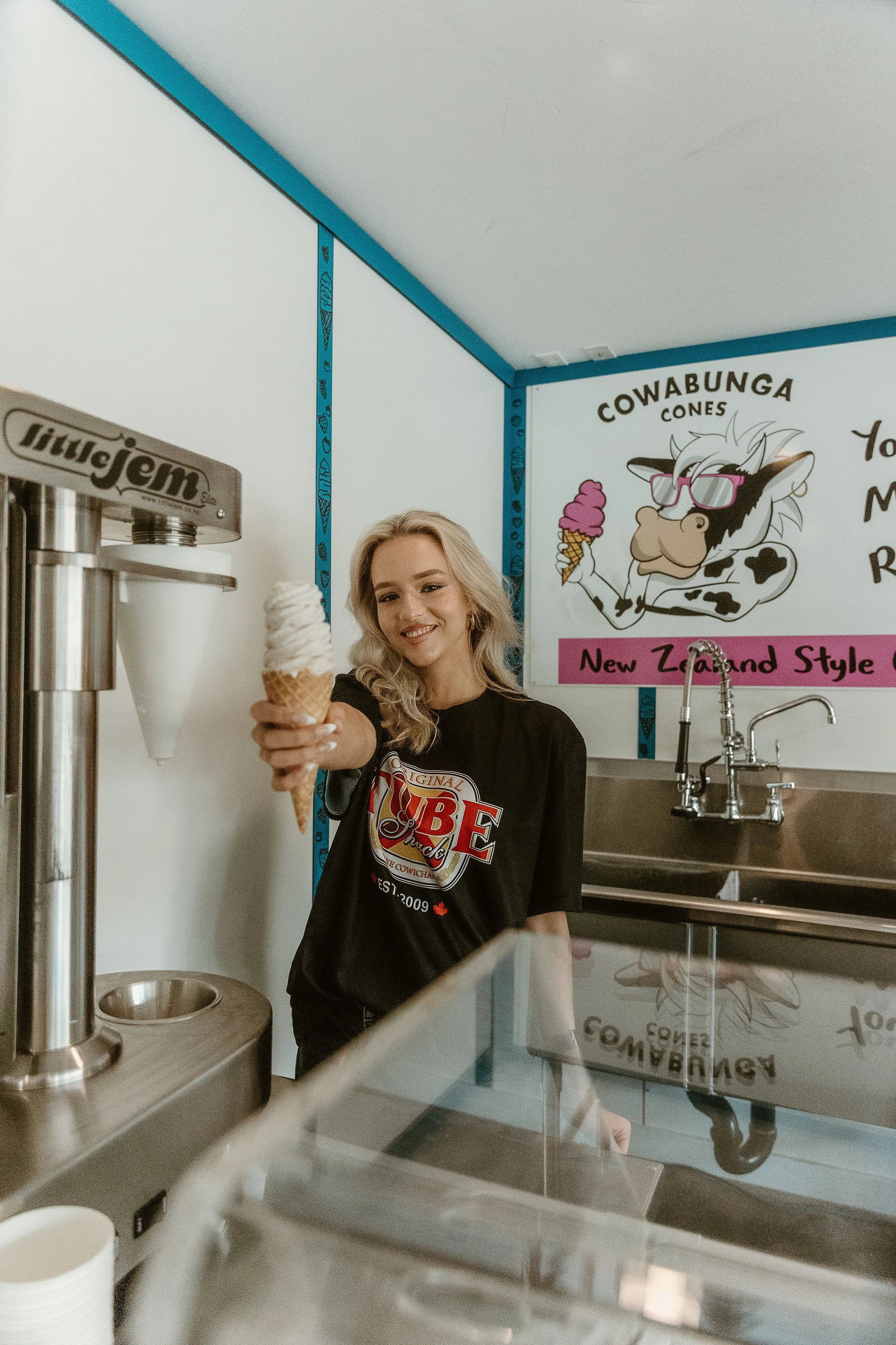A woman with blonde hair inside an ice cream shop, holding out an ice cream cone towards the camera and smiling. The shop features a sign with a cartoon cow and colorful decorations in the background.