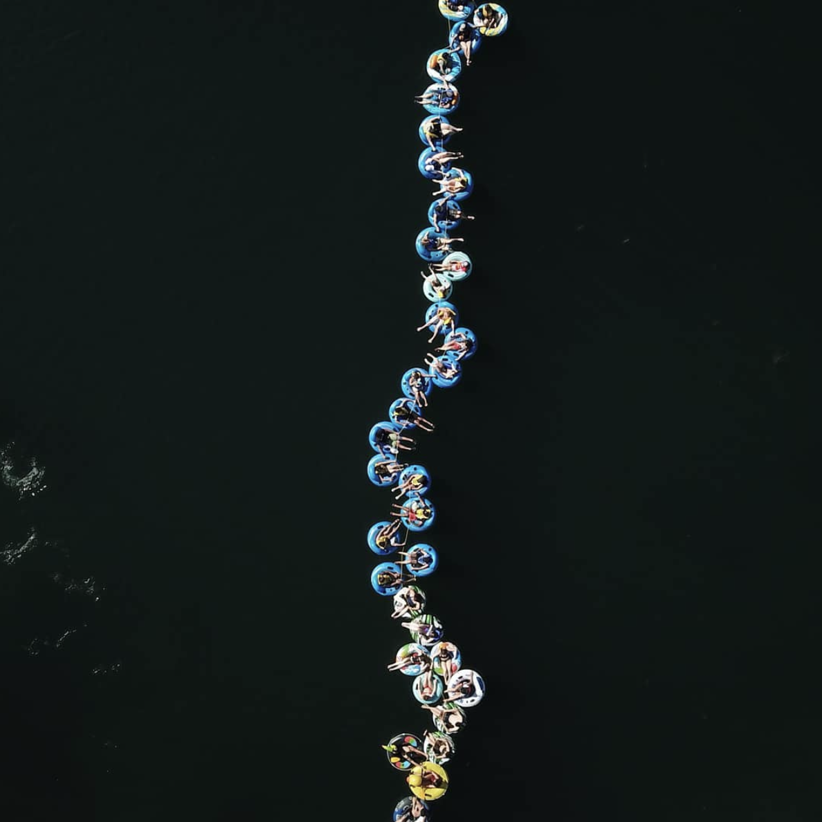 Aerial view of people on blue and yellow tubes arranged in a line on dark water.