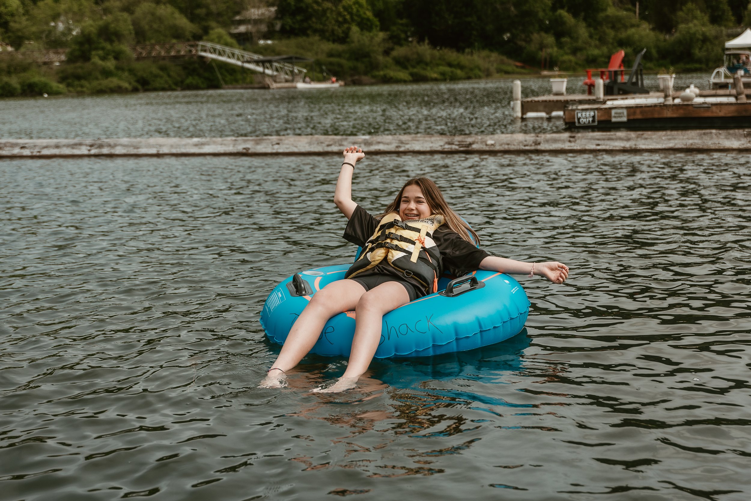 A young girl smiling and relaxing on a blue inflatable tube in the river, wearing a life jacket with her legs in the water.