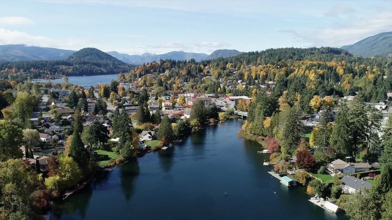 A scenic aerial view of a small town nestled along a river, with houses, trees, and mountains in the background during autumn.